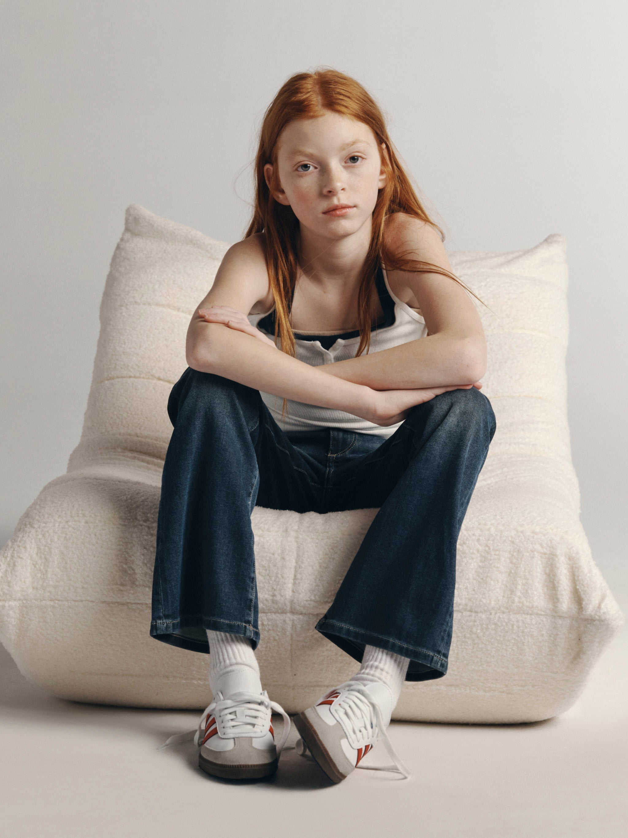 DL1961 Kids girls' Cleo bootcut jeans in Nightlight with a white tank, seated full-length on a cushion in a studio.
