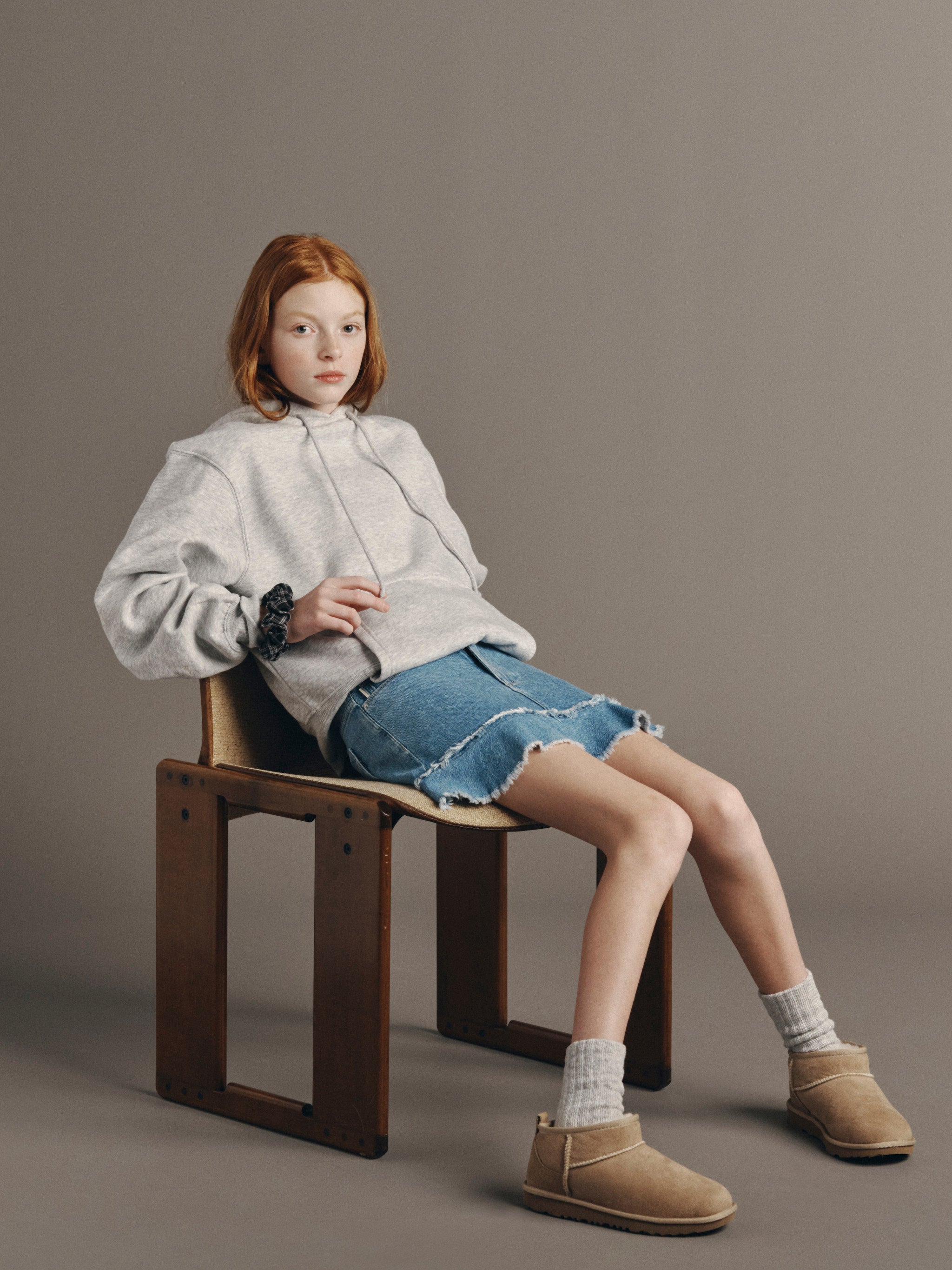 DL1961 Kids girls' Jenny ruffle denim skirt in Unraveled with a grey hoodie, seated on a chair in a studio.
