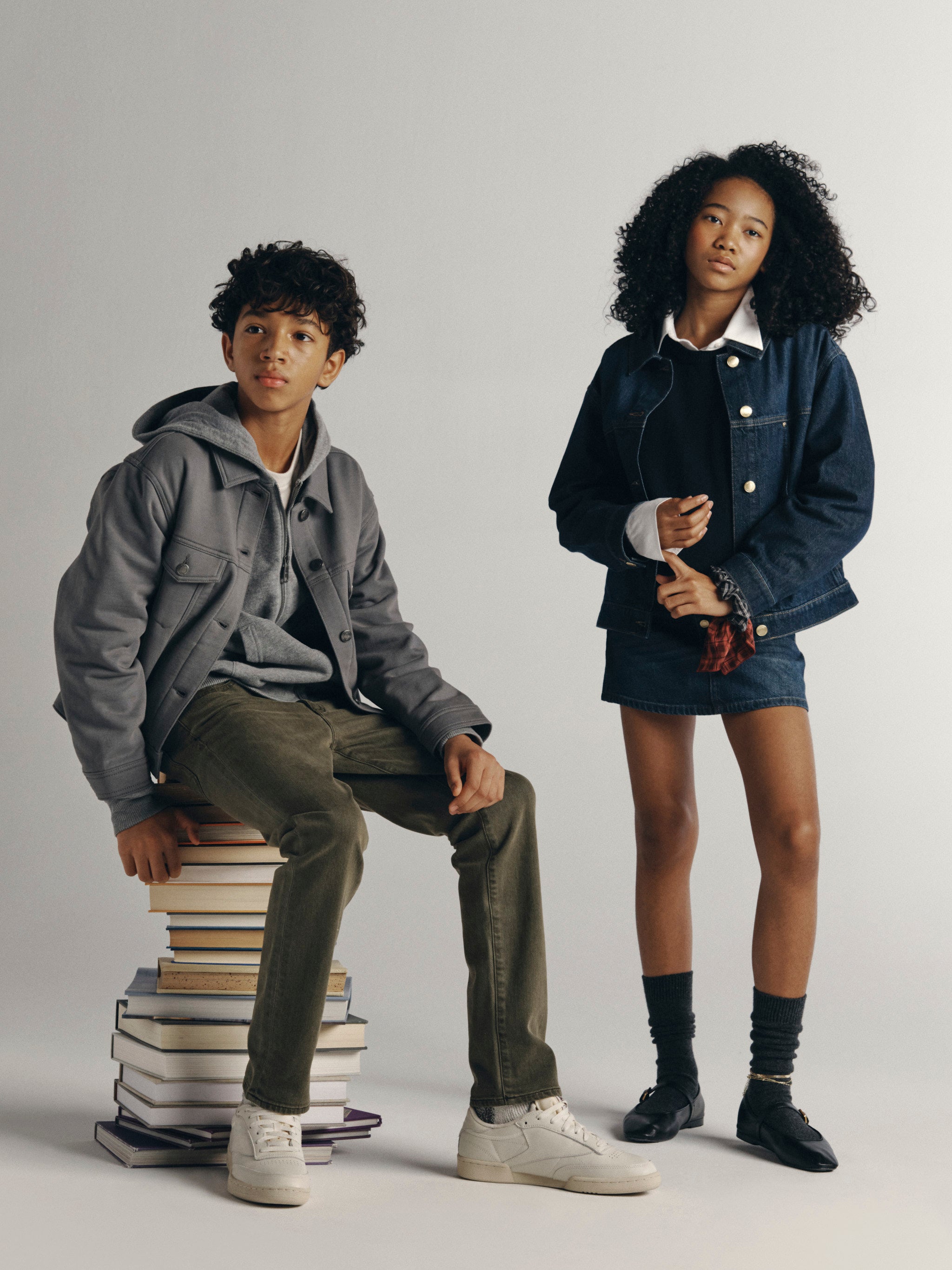 DL1961 Kids Fall 26 group shot — a boy in a grey Jordyn jacket and olive Brady jeans on a stack of books beside a girl in a denim Jenny skirt and jacket.