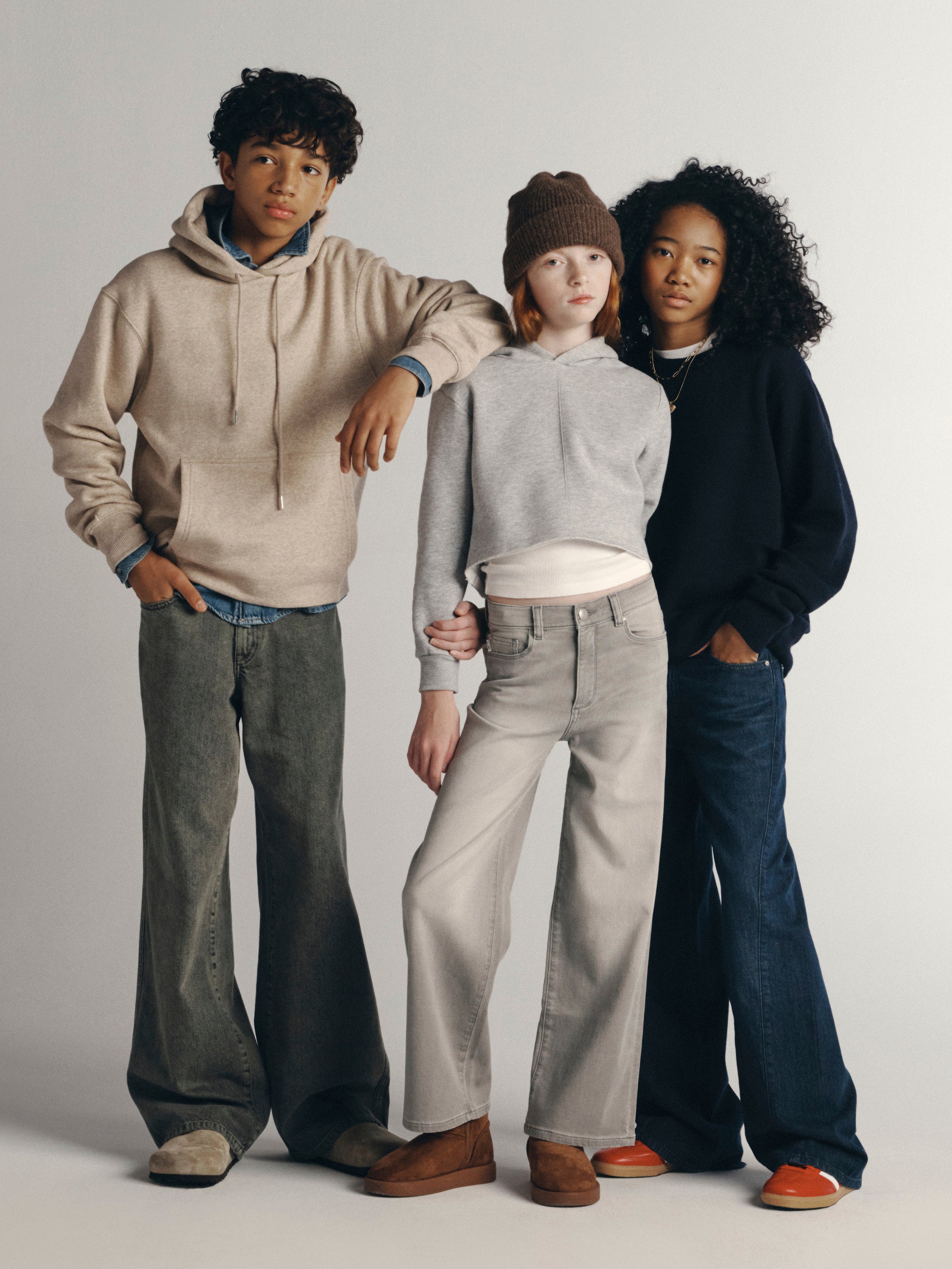 DL1961 Kids Fall 26 group shot — a boy in an oatmeal hoodie and grey jeans with two girls in a grey hoodie and wide-leg jeans, full-length studio.