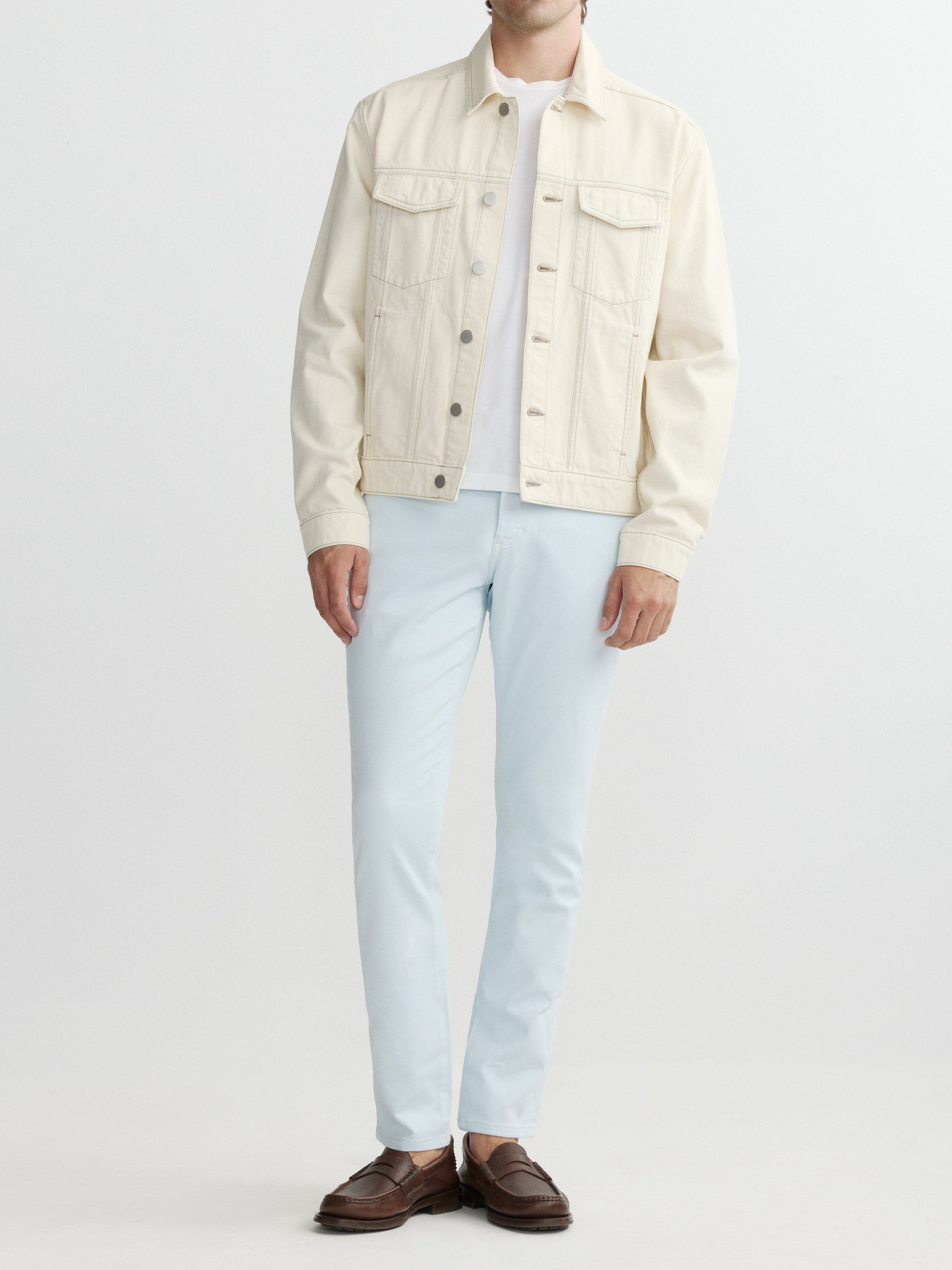 Person wearing a cream denim jacket, white shirt, light blue jeans, and brown shoes on a white background