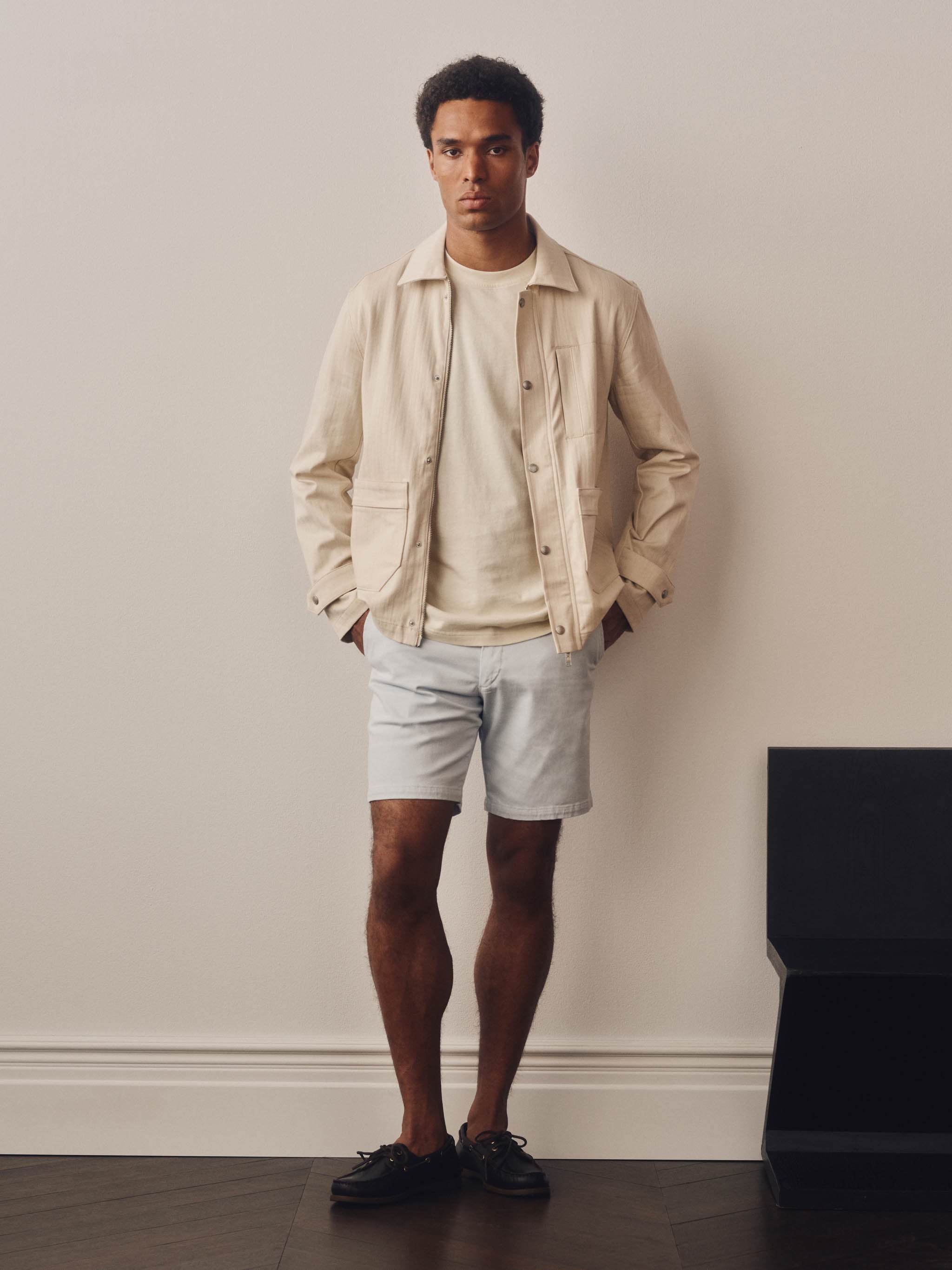 Man wearing a beige jacket and light blue shorts standing against a plain wall.