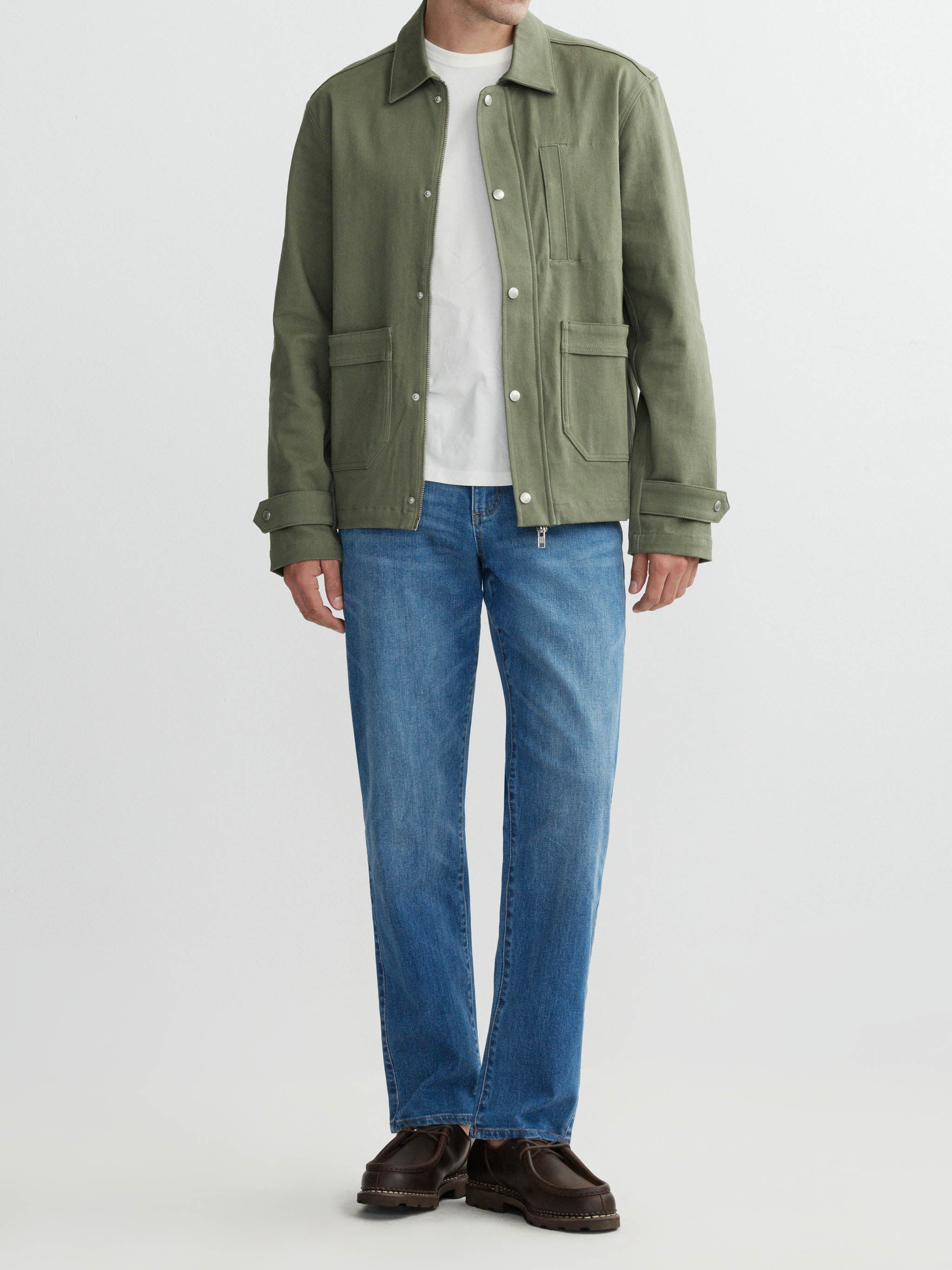 Person wearing a green jacket, white shirt, blue jeans, and brown shoes on a light gray background