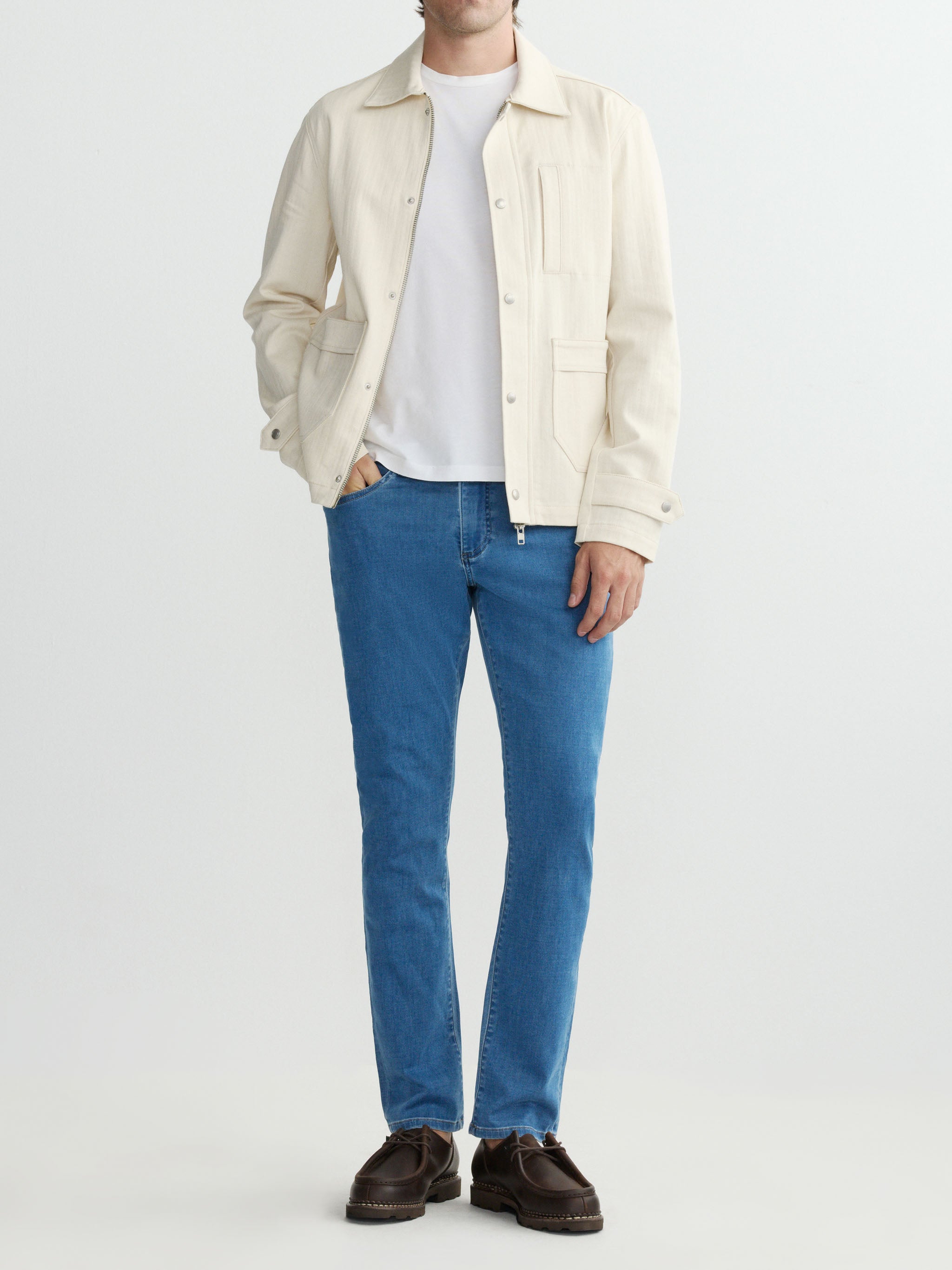 Person wearing a cream jacket, white shirt, blue jeans, and brown shoes on a light gray background
