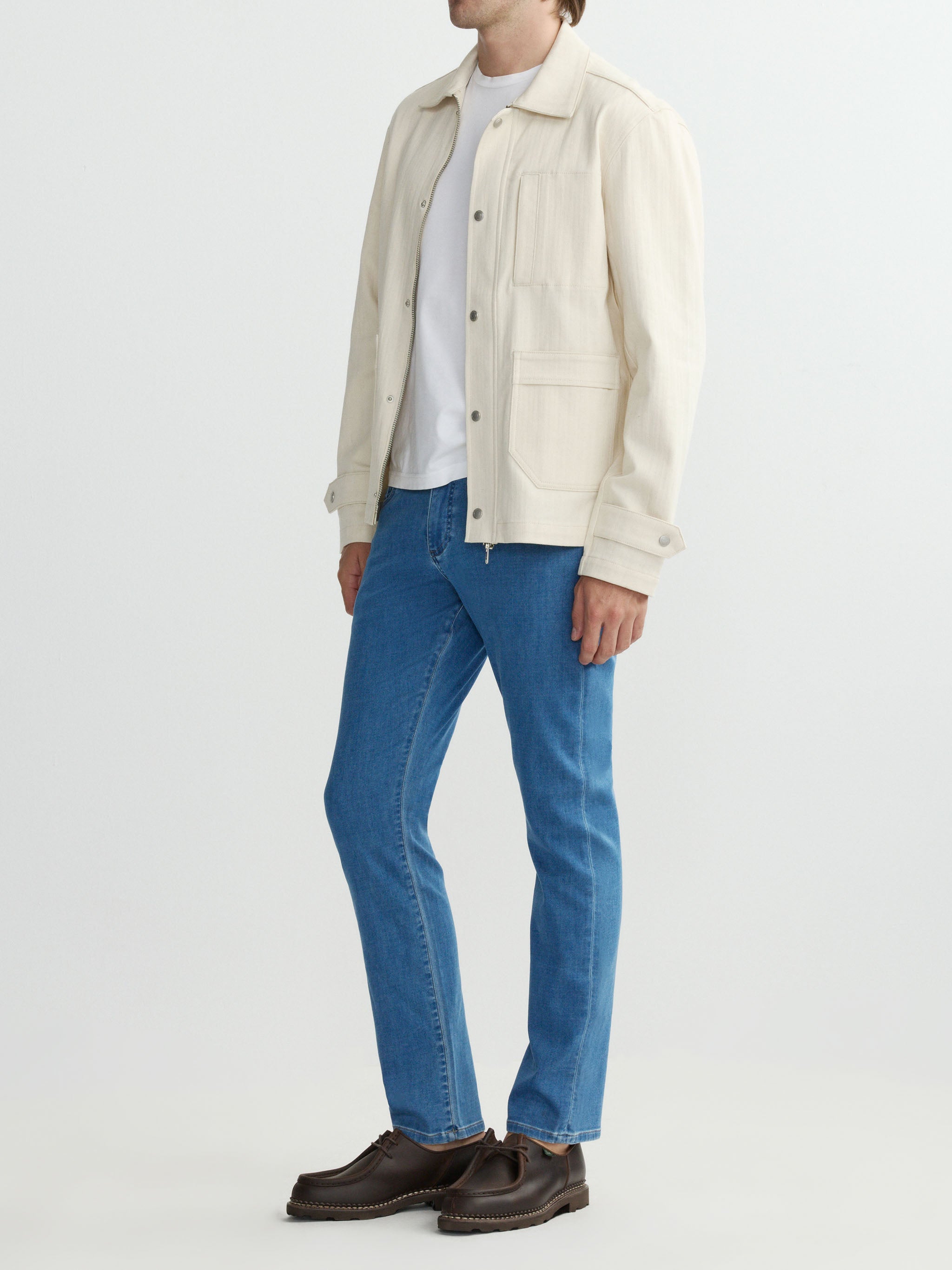 Person wearing a cream jacket, white shirt, blue jeans, and brown shoes on a white background