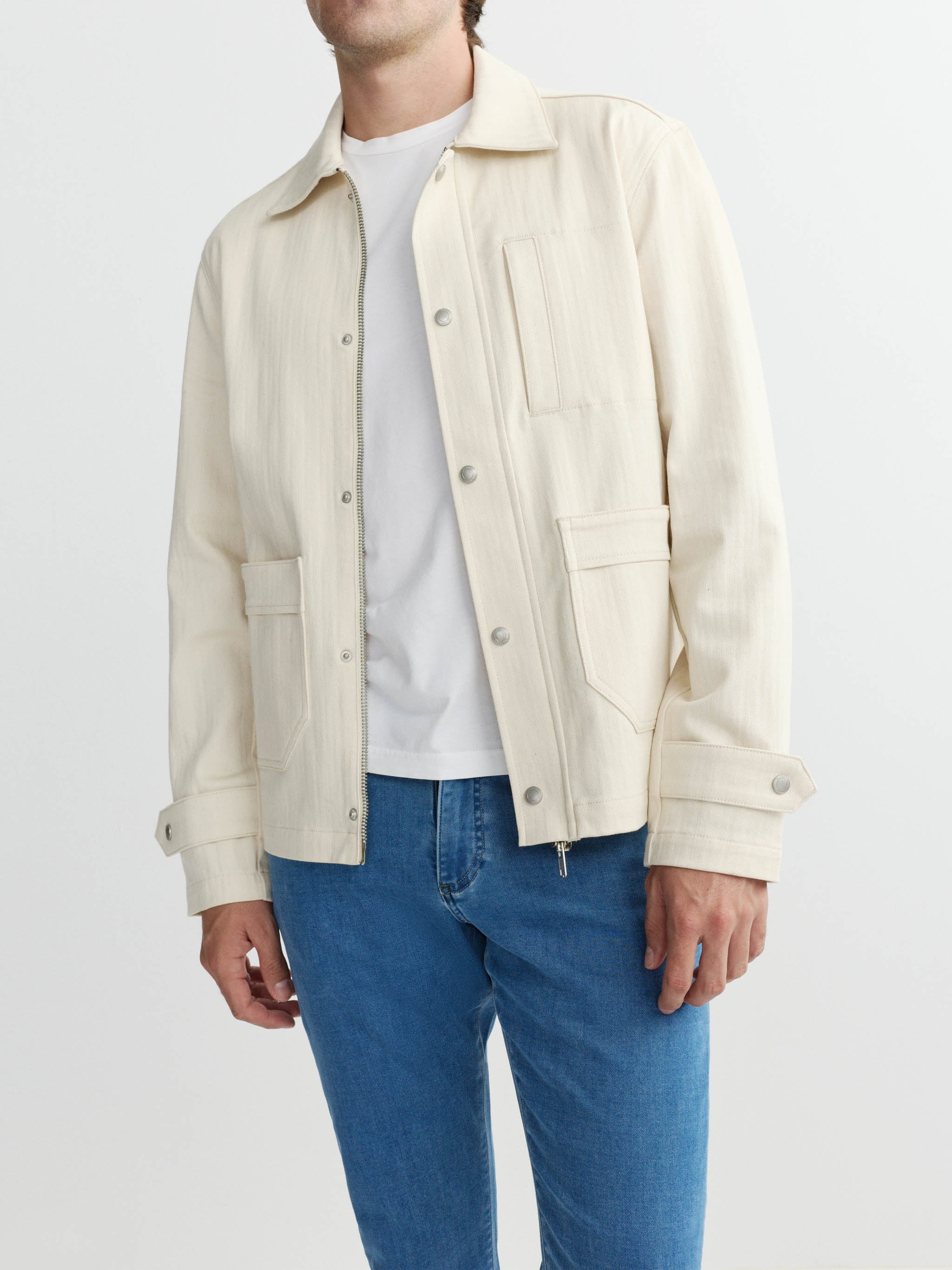 Person wearing a cream jacket over a white shirt and blue jeans on a light gray background