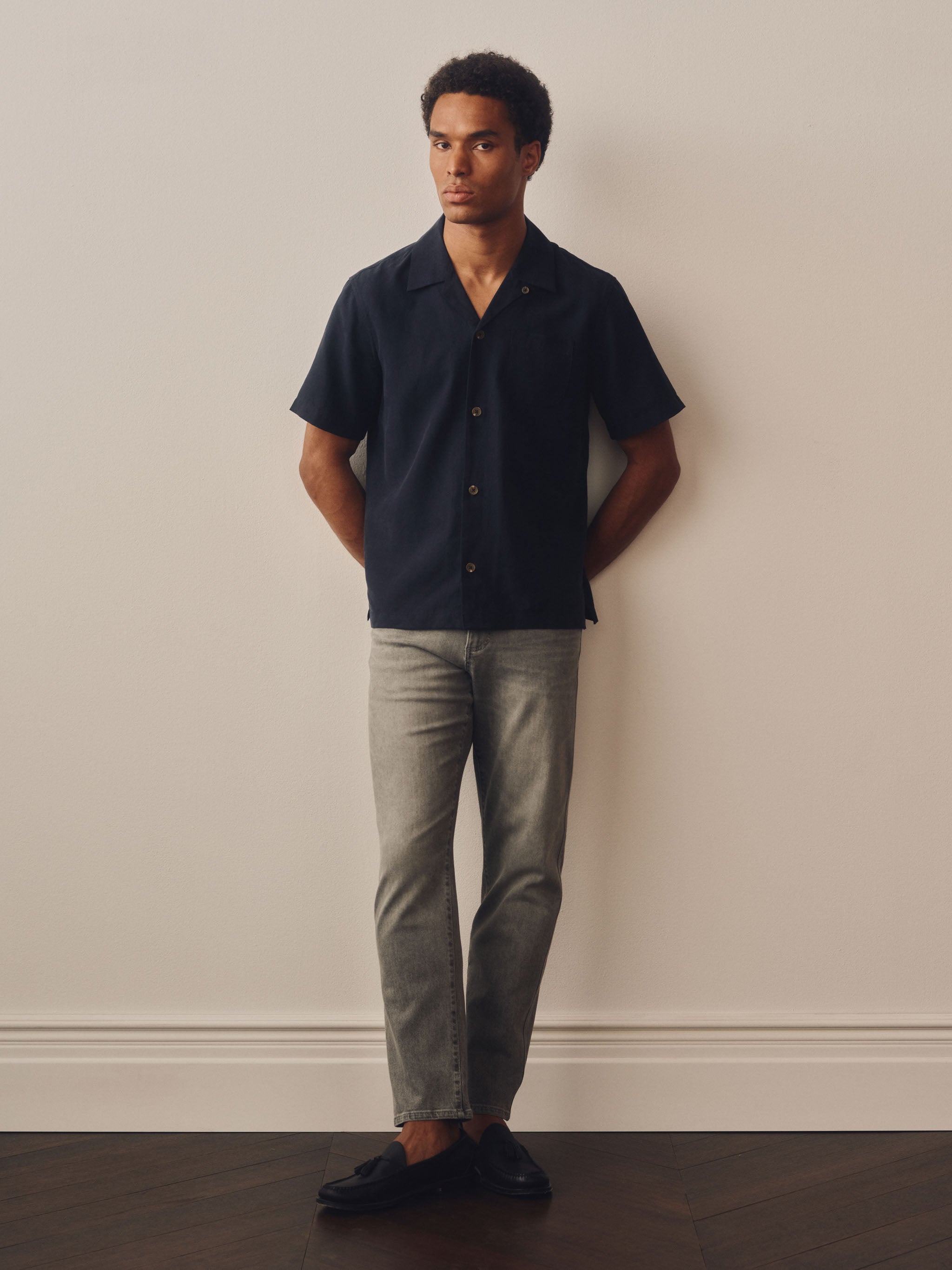 Man wearing a dark blue shirt and light gray pants against a plain wall.