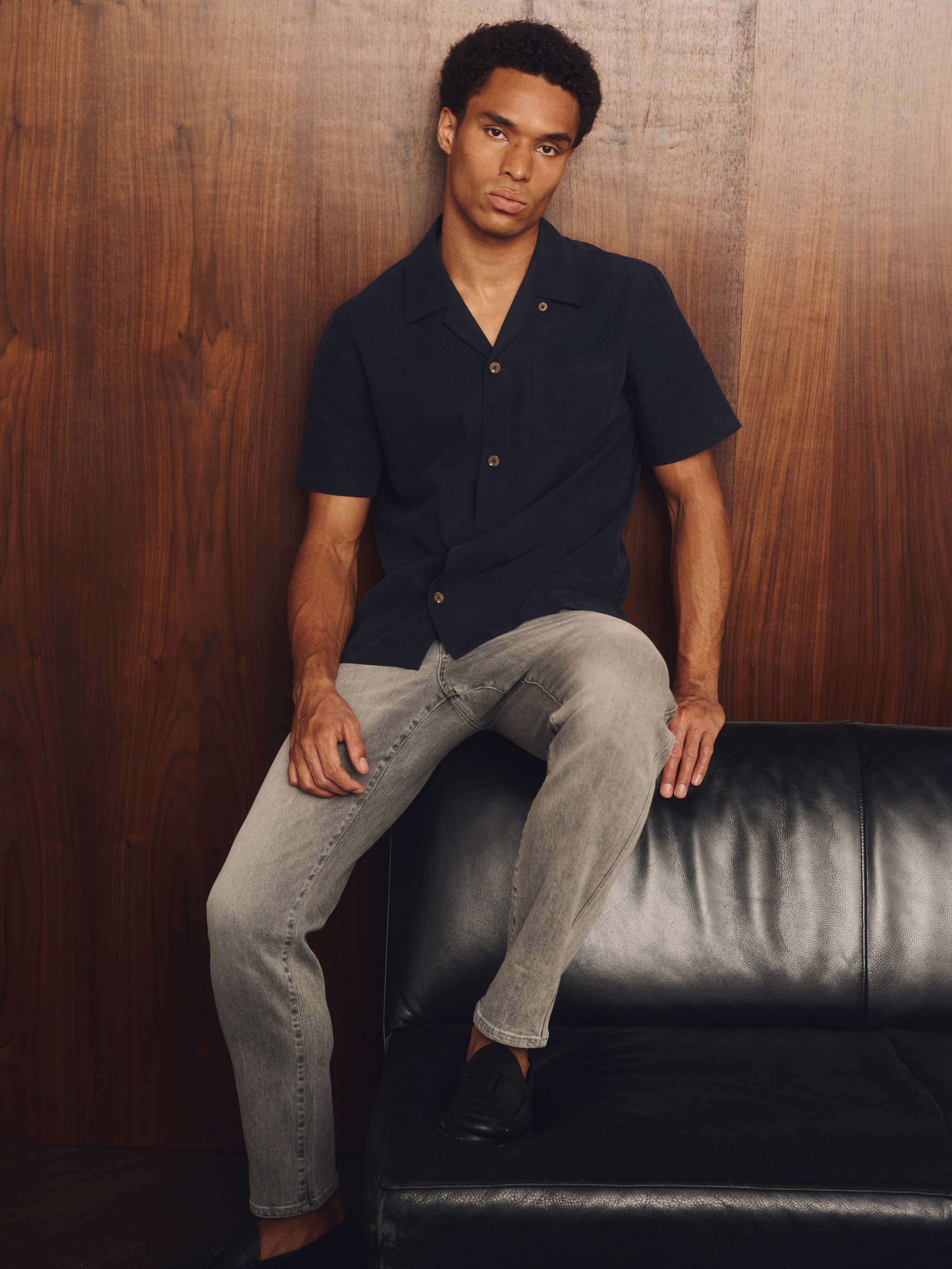 Man wearing a navy short-sleeve shirt with grey pants on a wood background