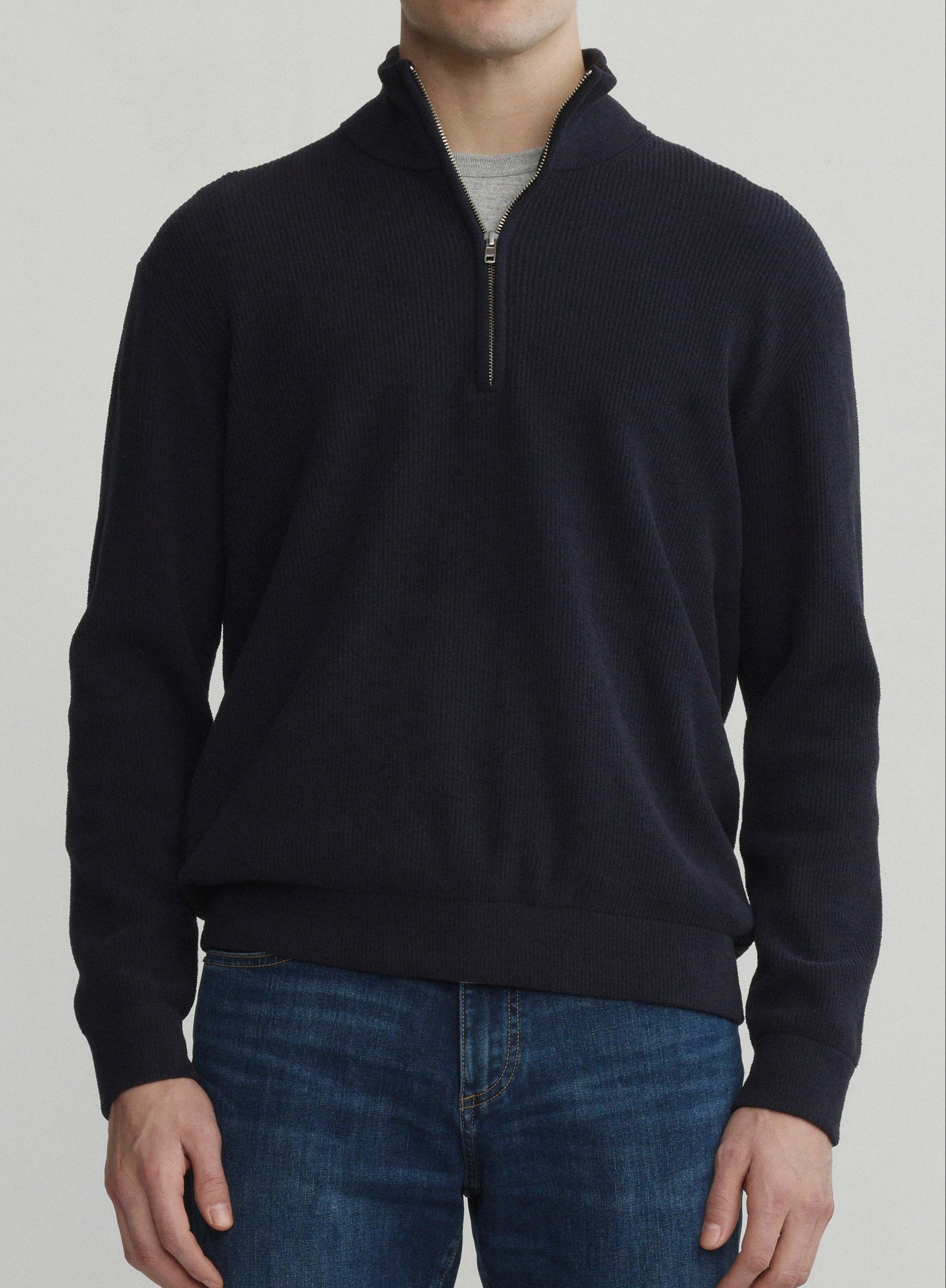 A man wearing a dark navy quarter-zip sweater, blue jeans, and brown shoes, standing against a light gray studio background.