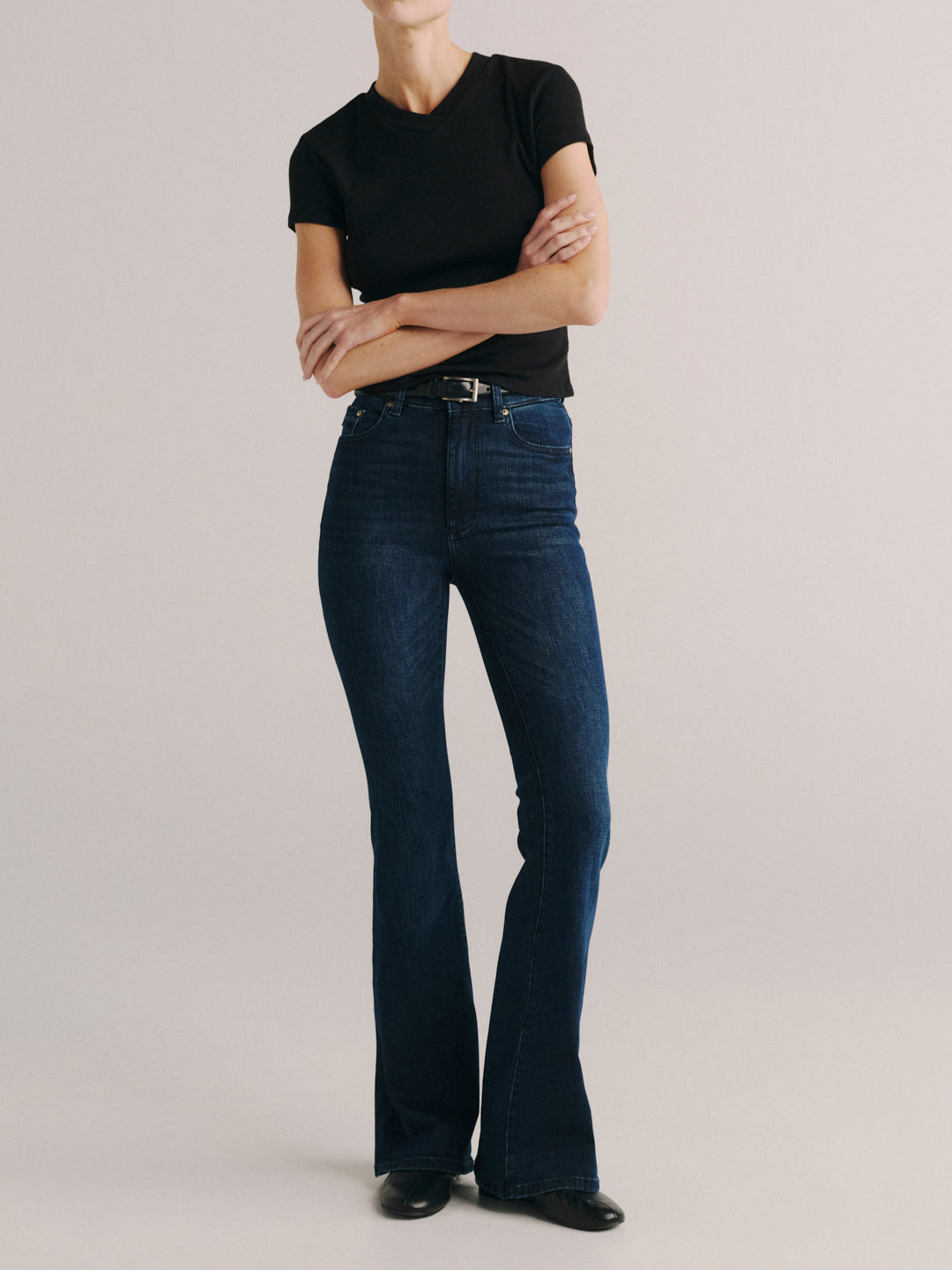 A model wearing dark blue high-waisted flare jeans with a black t-shirt and black shoes, standing against a light gray studio background.