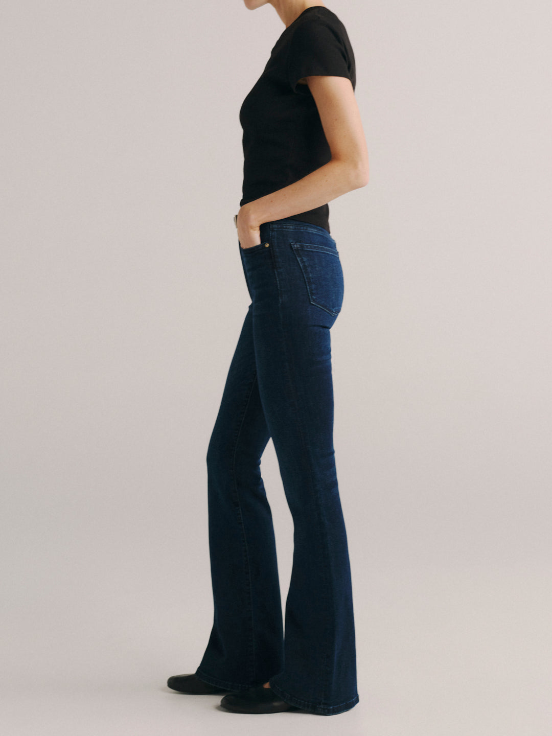 A model shown from the side wearing dark blue flared jeans, a black short-sleeve top, and black shoes against a plain light gray studio background.