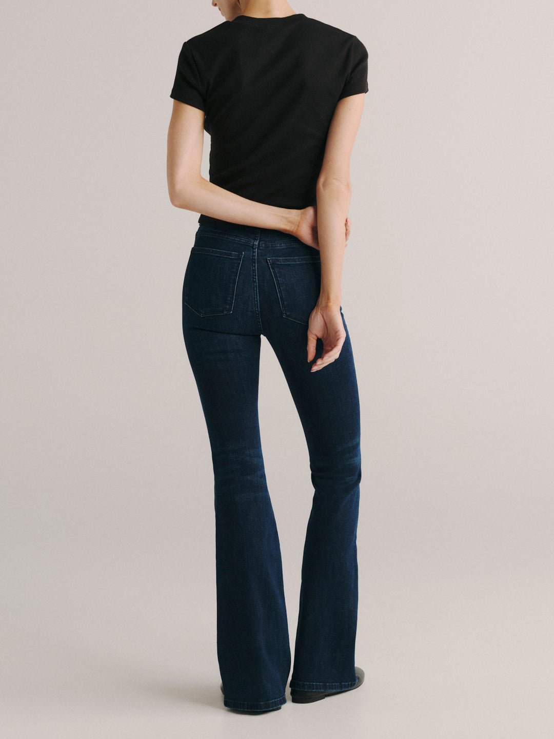 A model shown from the back wearing dark blue flare jeans and a black t-shirt, standing against a neutral light gray studio background.