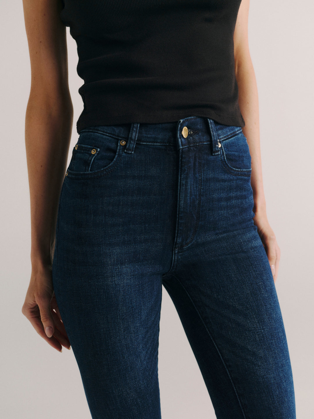 A close-up of a model wearing dark blue denim jeans with a black sleeveless top, shown from the chest down against a light gray studio background.