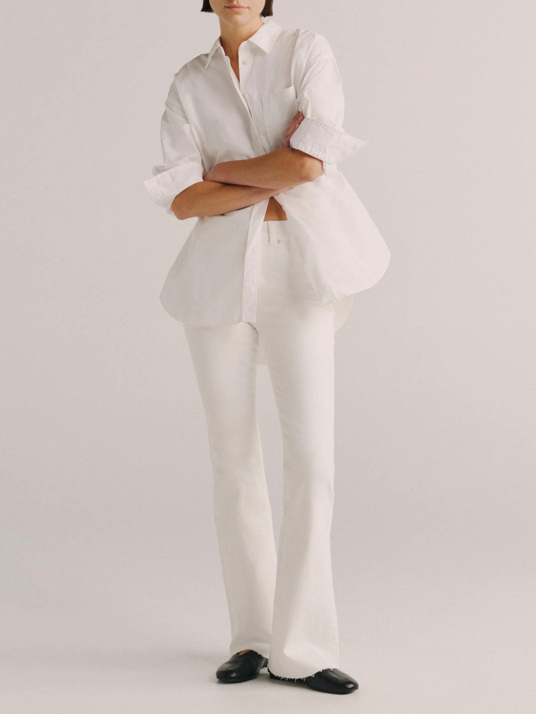 A model wearing a white long-sleeve button-up shirt with rolled cuffs, white trousers, and black shoes, standing against a light gray studio background.