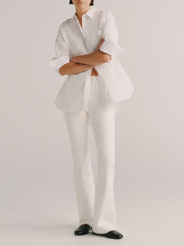 A model wearing a white long-sleeve button-up shirt with rolled cuffs, white trousers, and black shoes, standing against a light gray studio background.