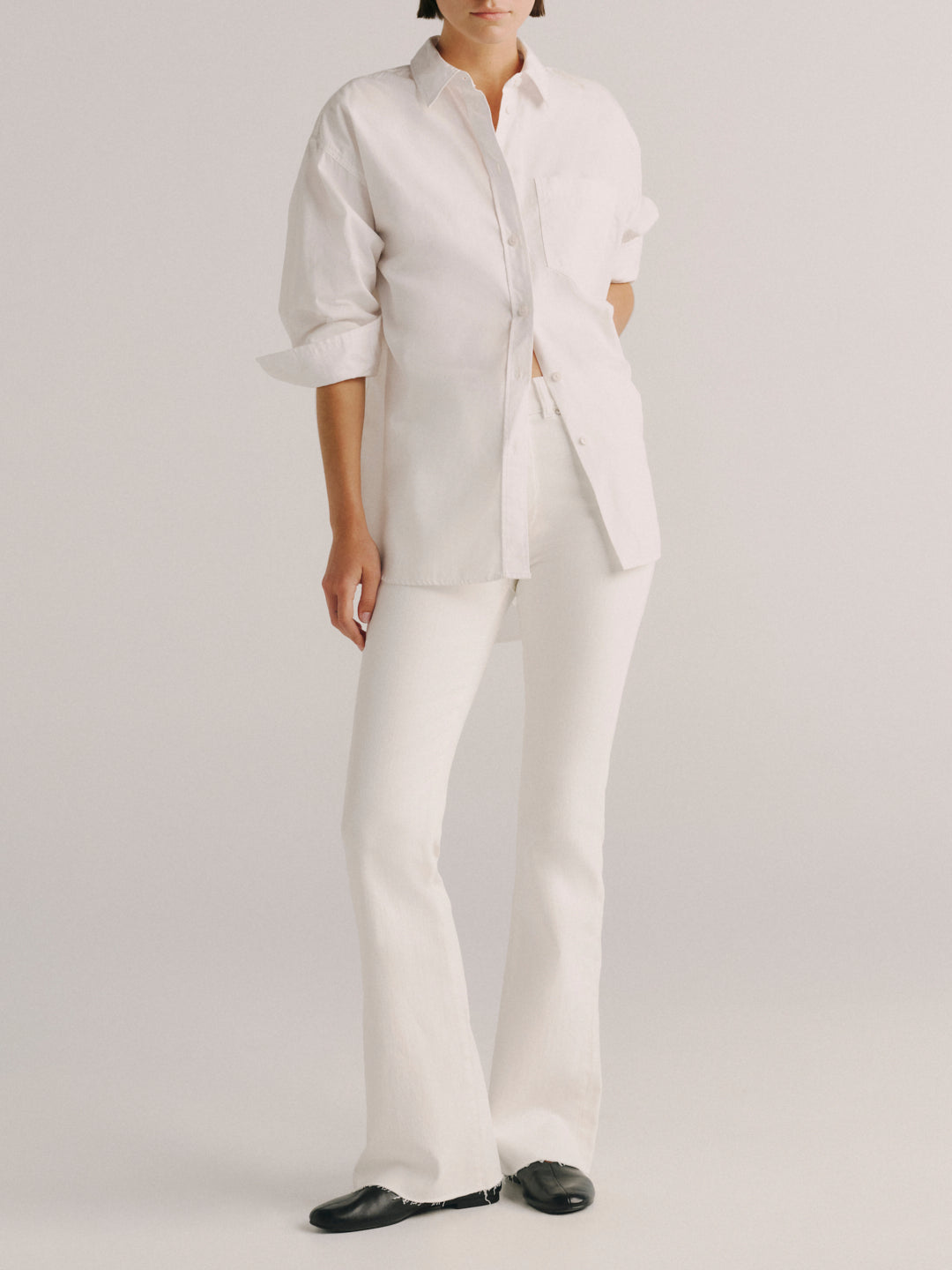 A model wearing a white long-sleeve button-up shirt and matching white wide-leg trousers, paired with black shoes, against a light gray studio background.