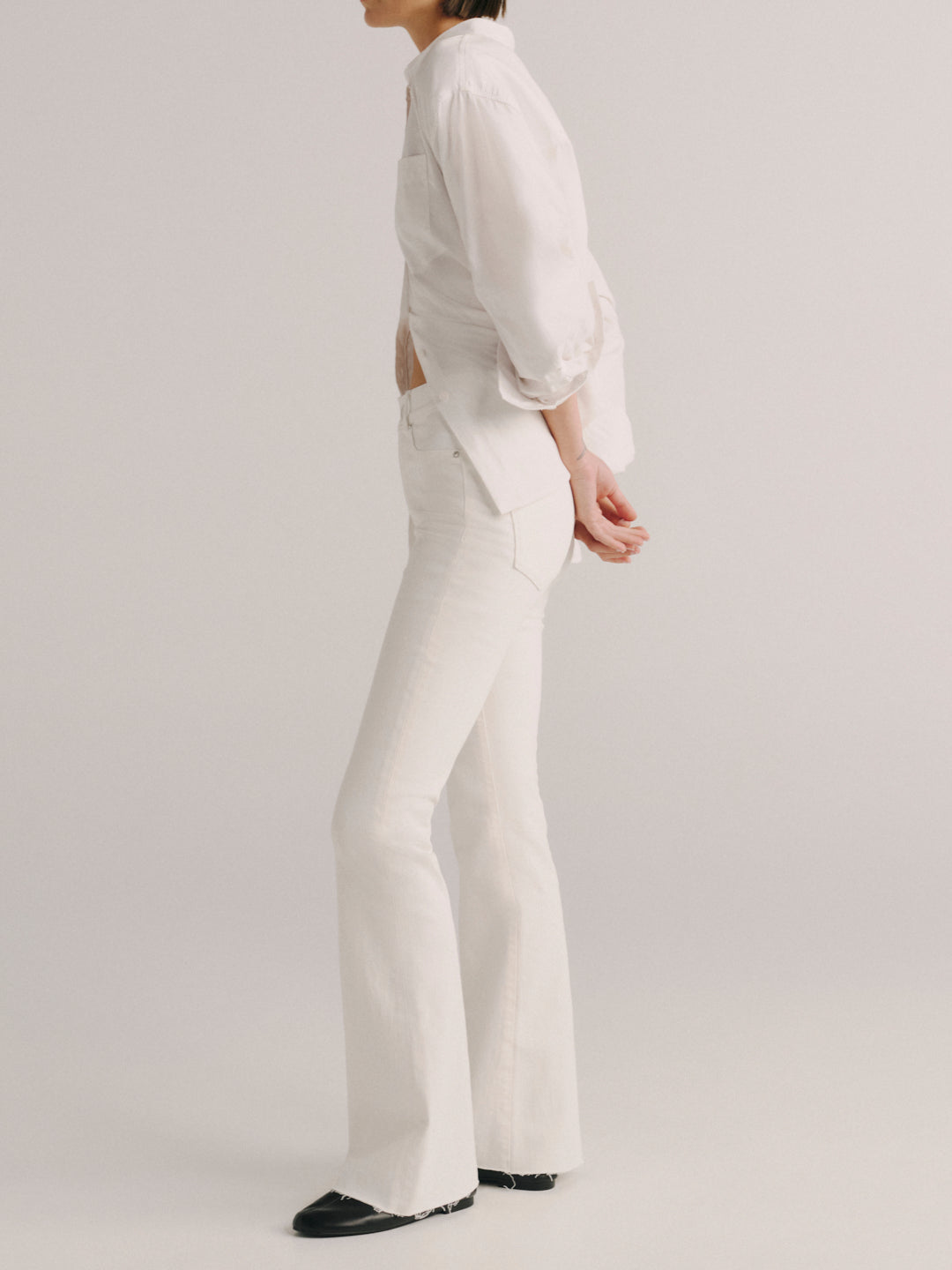 A model shown from the side wearing a white long-sleeve blouse with ruffled cuffs and white flared trousers, paired with black shoes, against a light gray studio background.