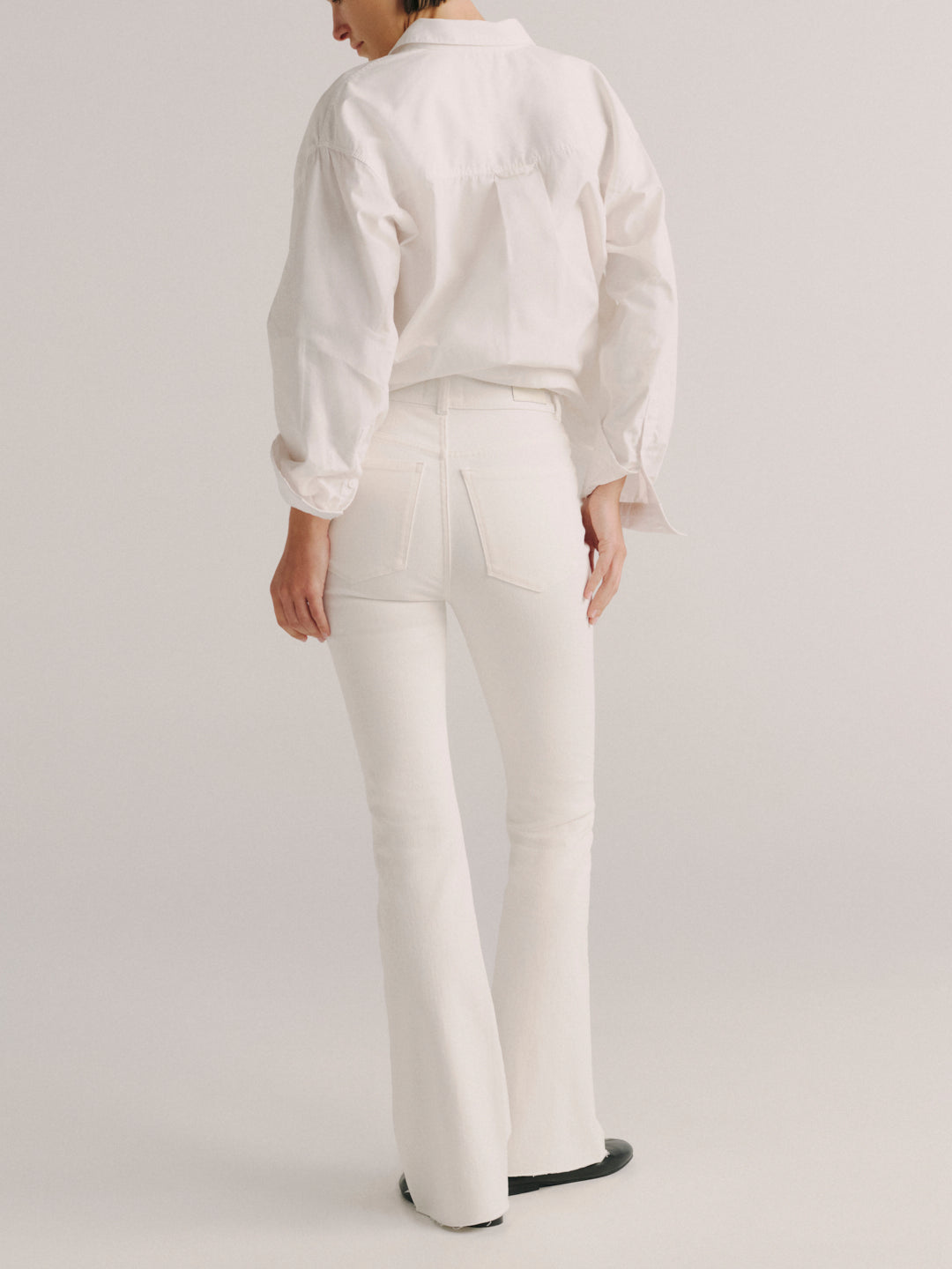 A model shown from the back wearing a white long-sleeve button-up shirt and matching white flared jeans, paired with black shoes, against a light gray studio background.