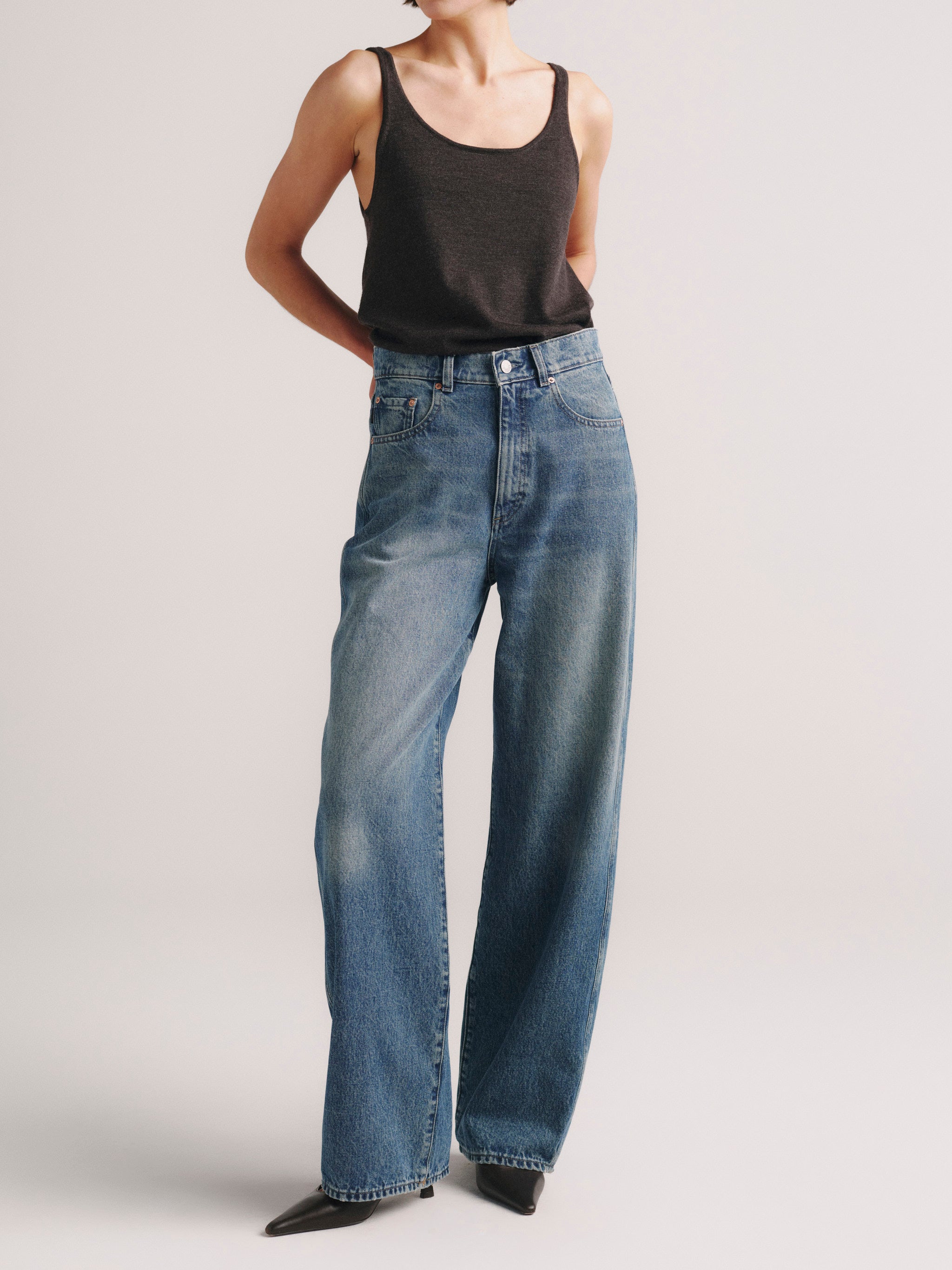 A model wearing a pair of high-waisted, wide-leg blue denim jeans with a faded wash, paired with a black tank top and black pointed-toe heels, photographed against a light gray studio background.