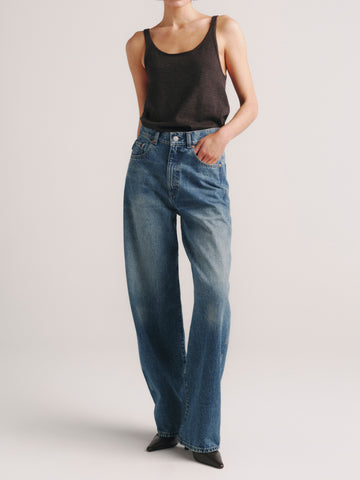 A model wearing a pair of high-waisted, wide-leg blue denim jeans with a black tank top, standing against a light gray studio background.