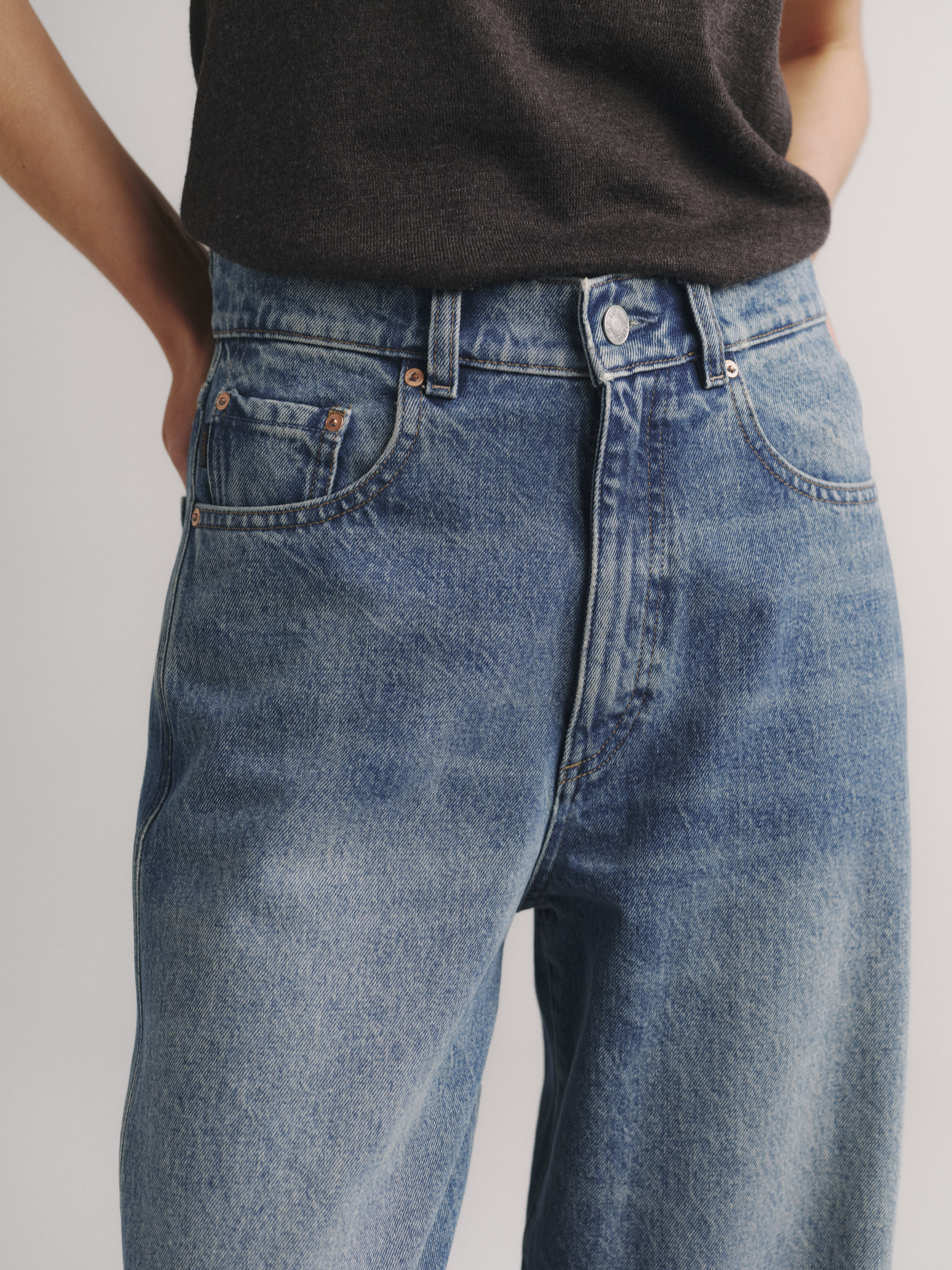 Close-up of a person wearing high-waisted, wide-leg blue denim jeans with a dark charcoal t-shirt, shown from the waist down against a light gray background.