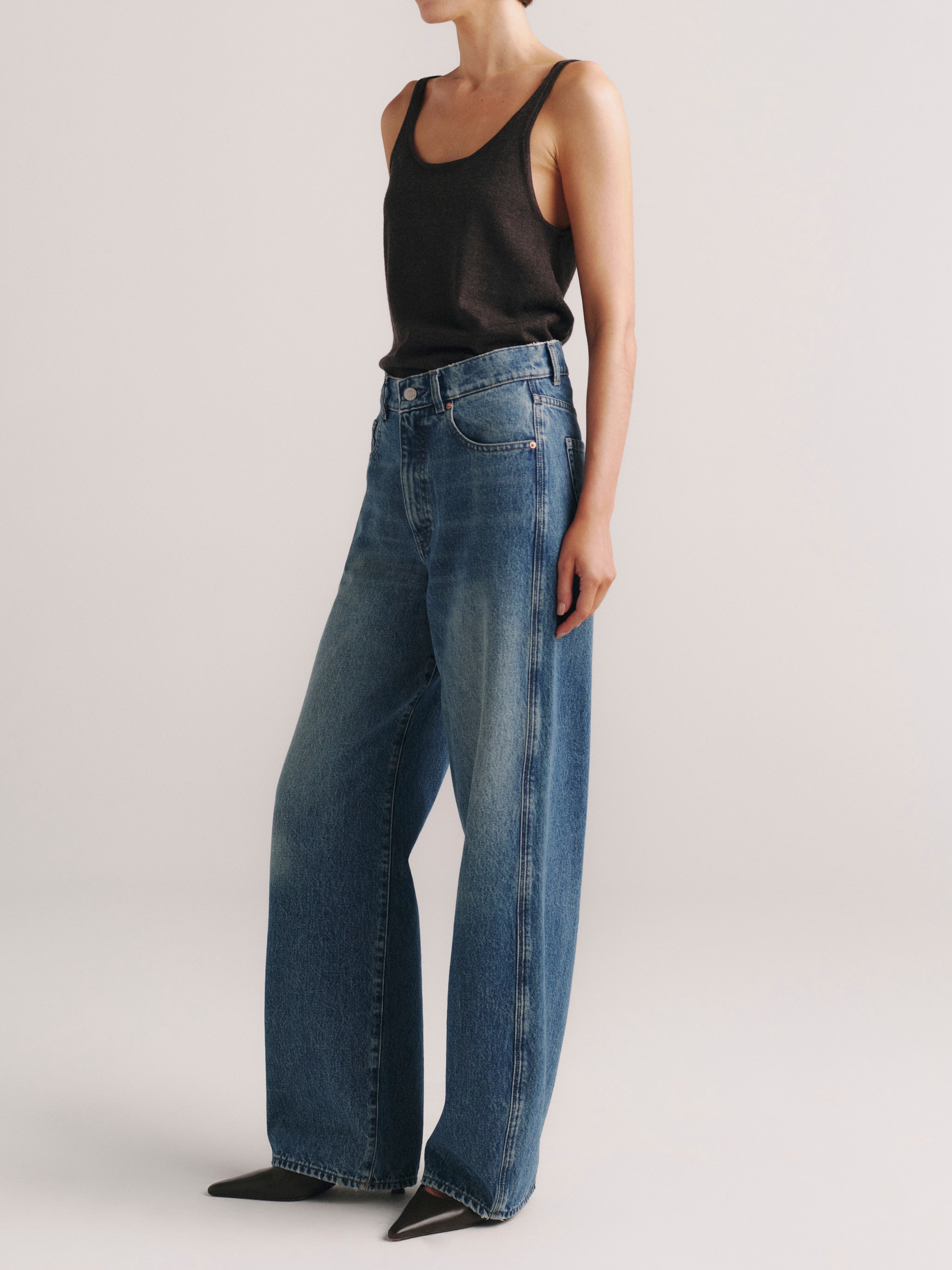 A model wearing a pair of high-waisted, wide-leg blue denim jeans with a black tank top, shown from the neck down against a light gray studio background.