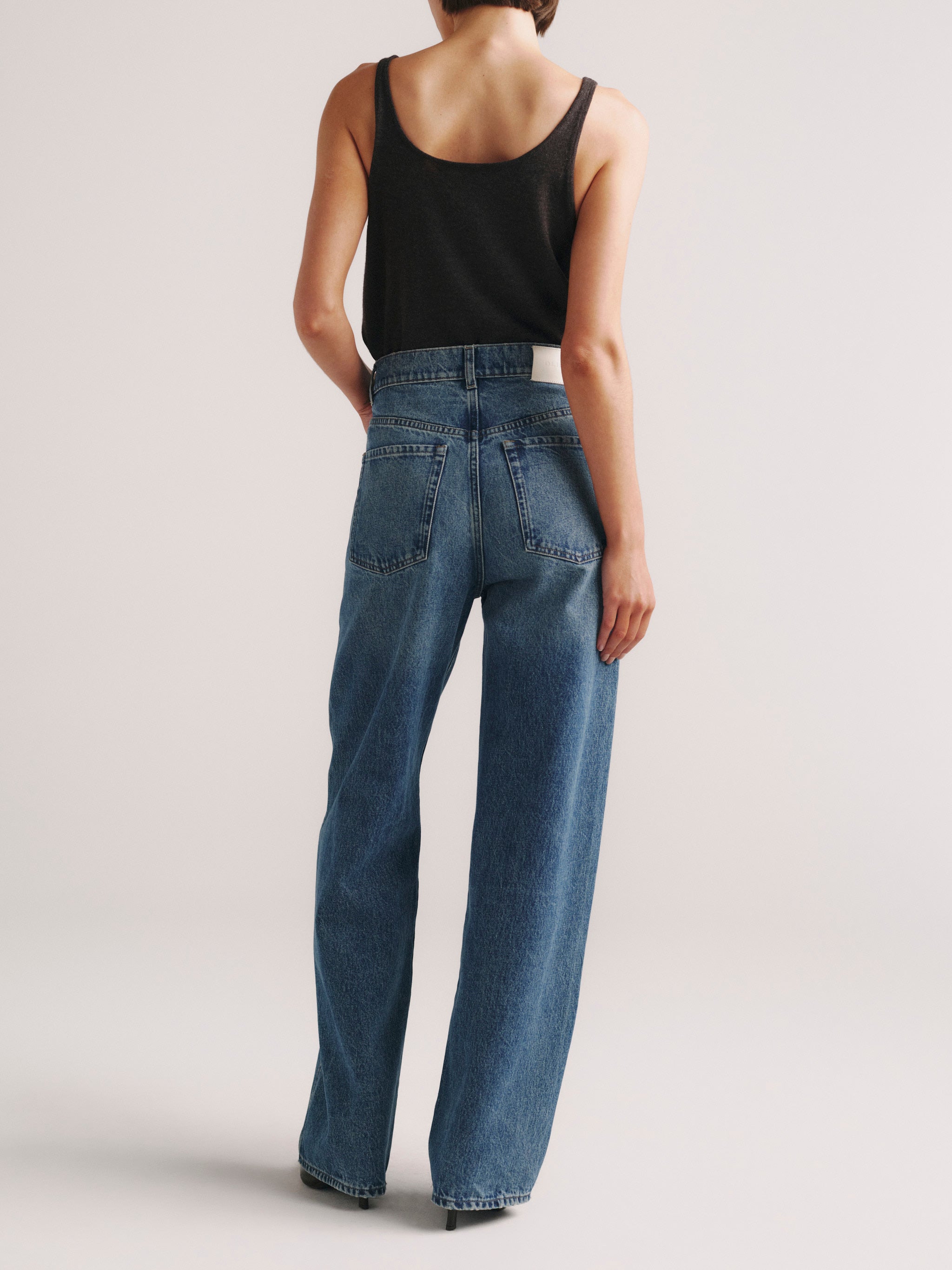 A model shown from the back wearing high-waisted, wide-leg blue jeans and a black tank top against a light gray studio background.