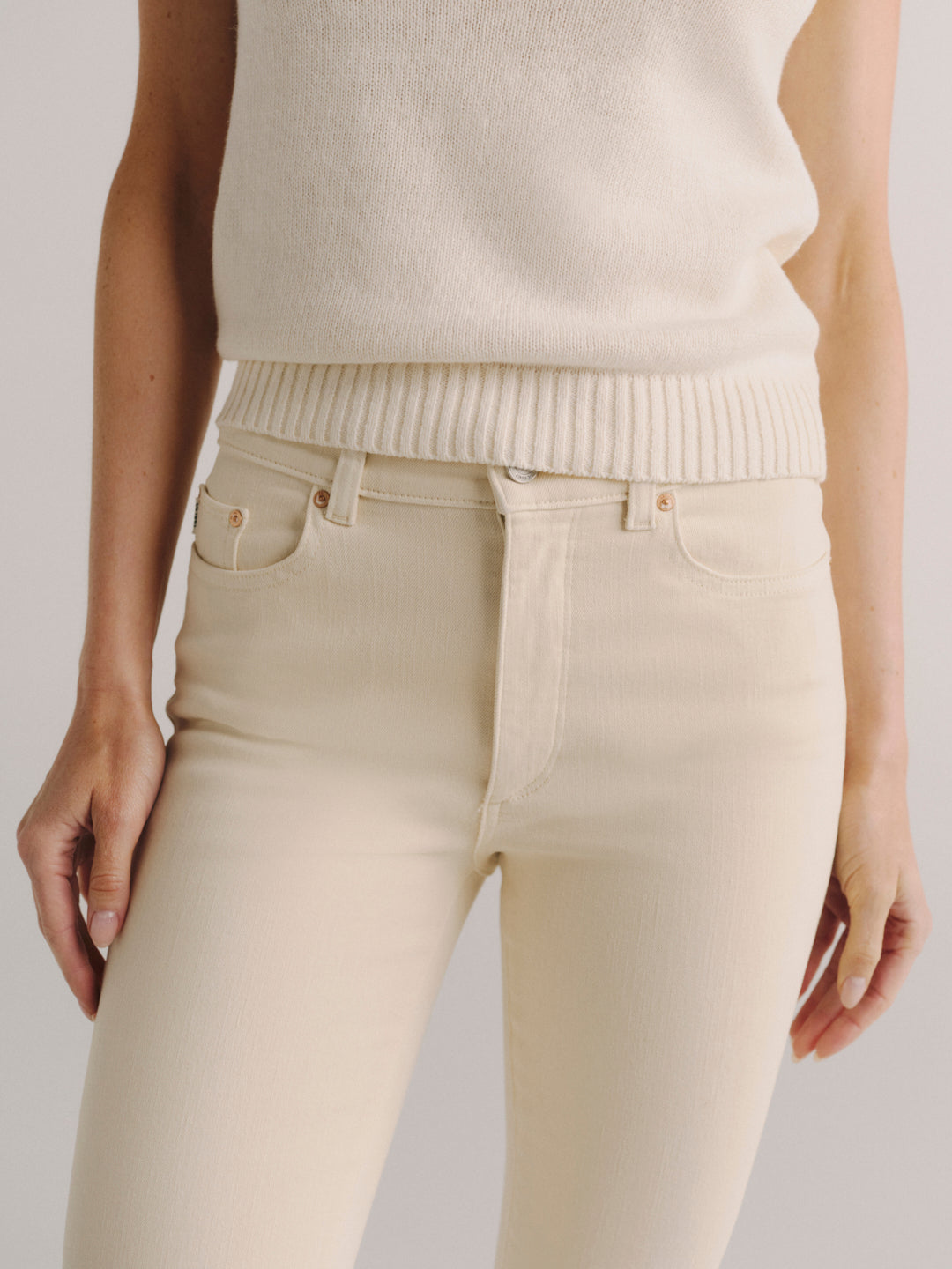 A person wearing cream-colored ribbed knit sweater vest and matching cream skinny jeans against a light gray studio background.