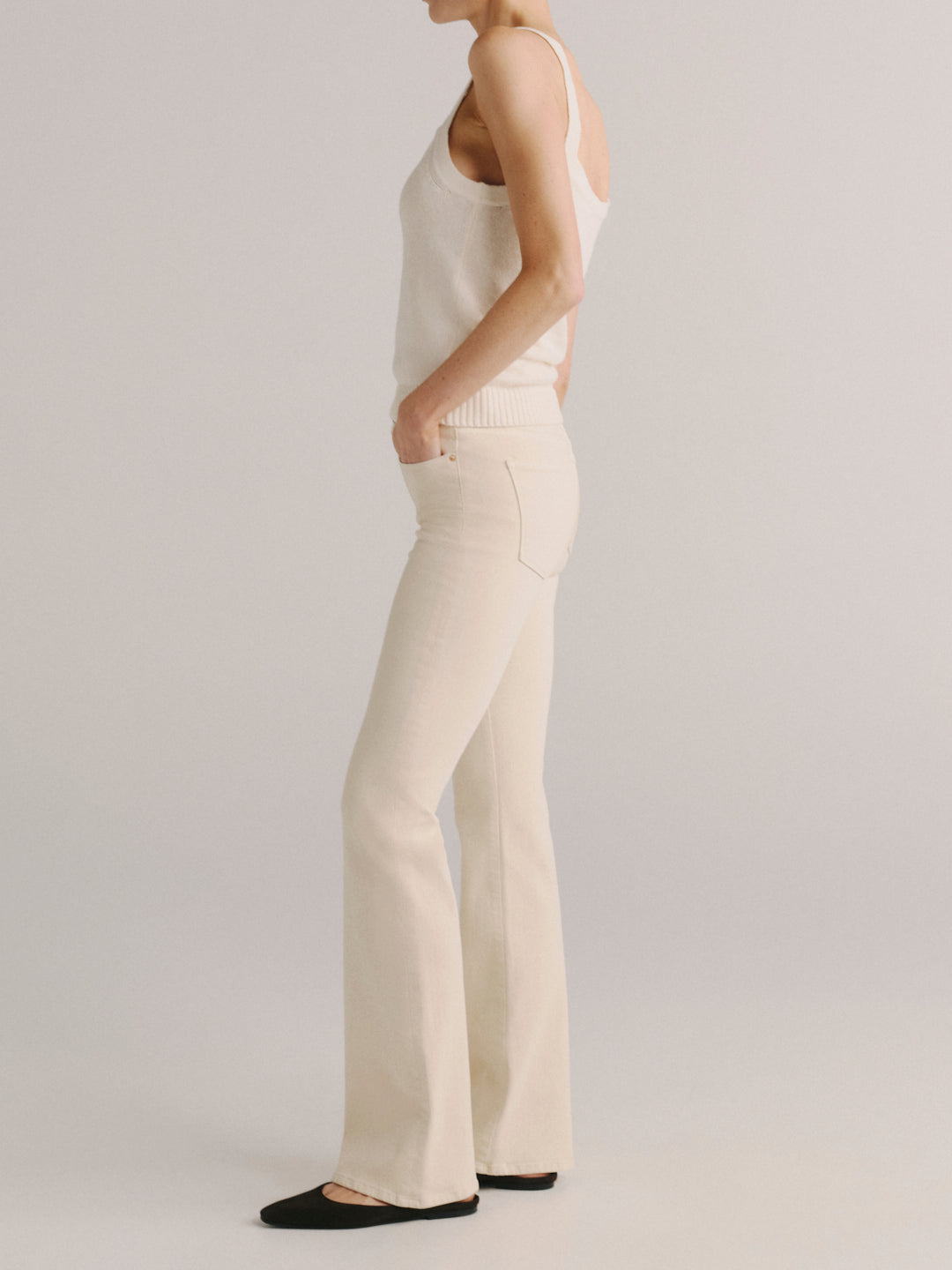 A model shown from the side wearing cream-colored flared pants and a matching cream knit tank top, paired with black flats, against a light gray studio background.
