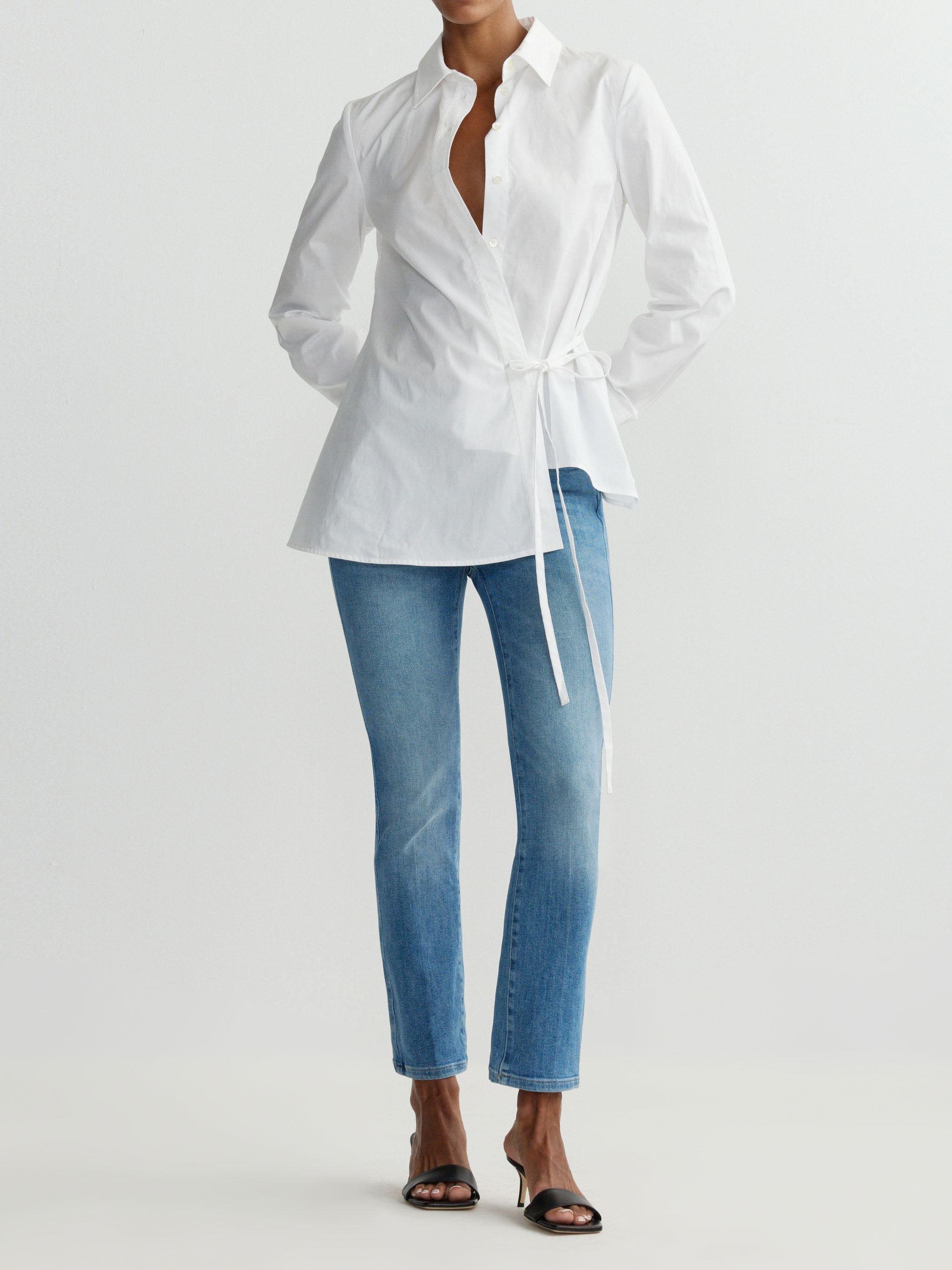 Person wearing a white blouse with a tie and blue jeans on a light gray background