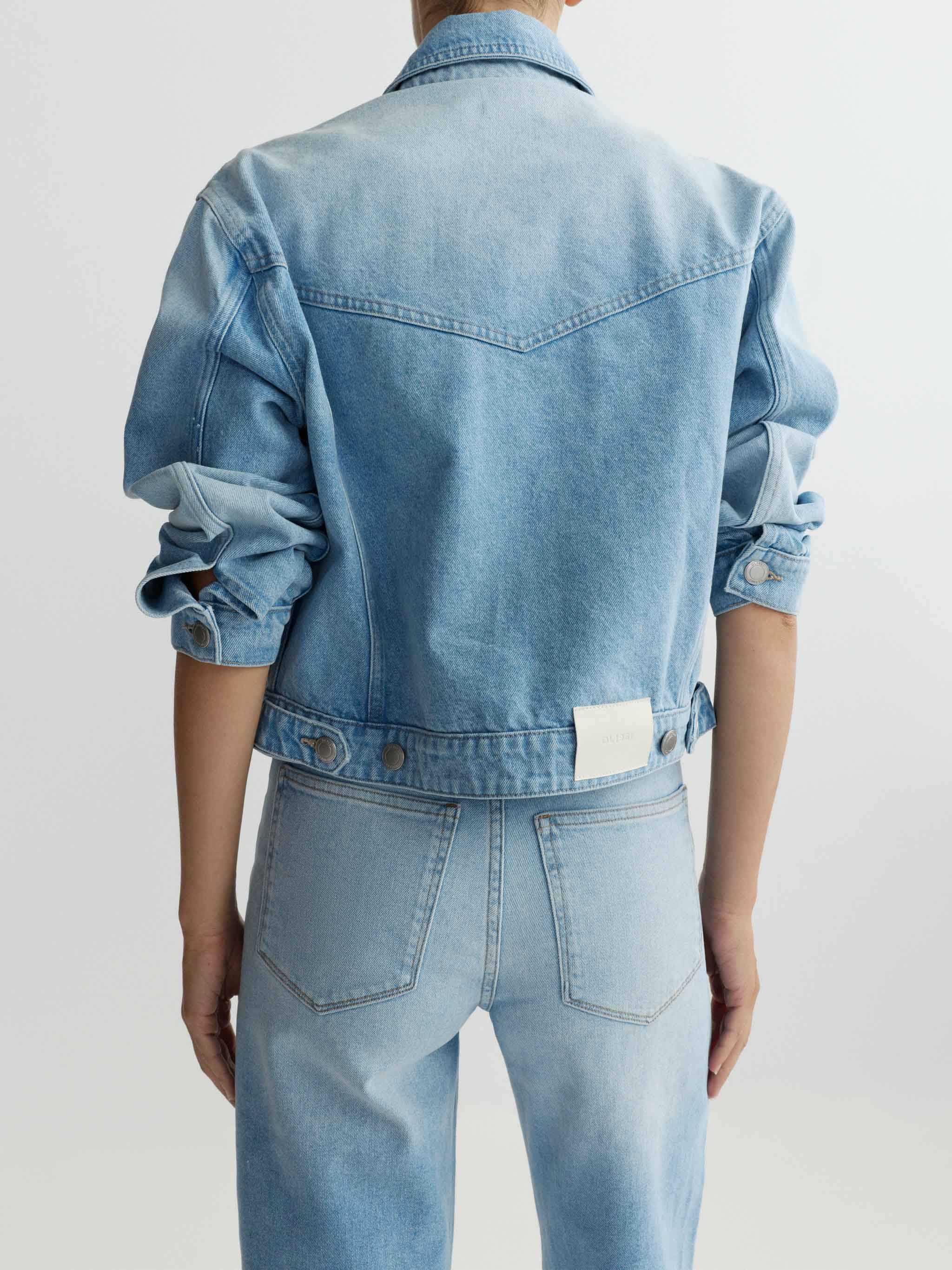 Person wearing a light blue denim jacket and jeans on a plain background