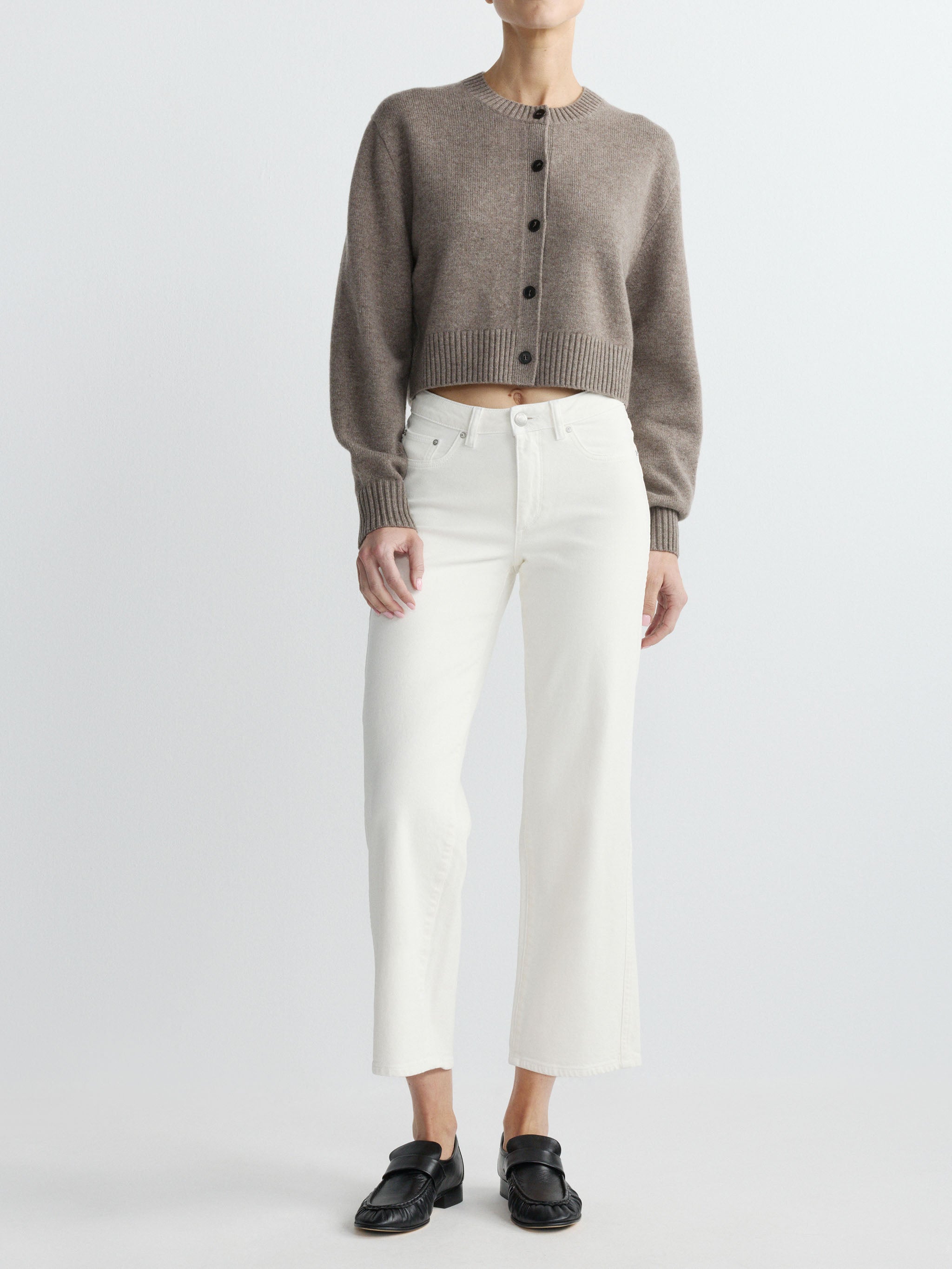 Person wearing a brown cardigan, white pants, and black shoes on a light gray background