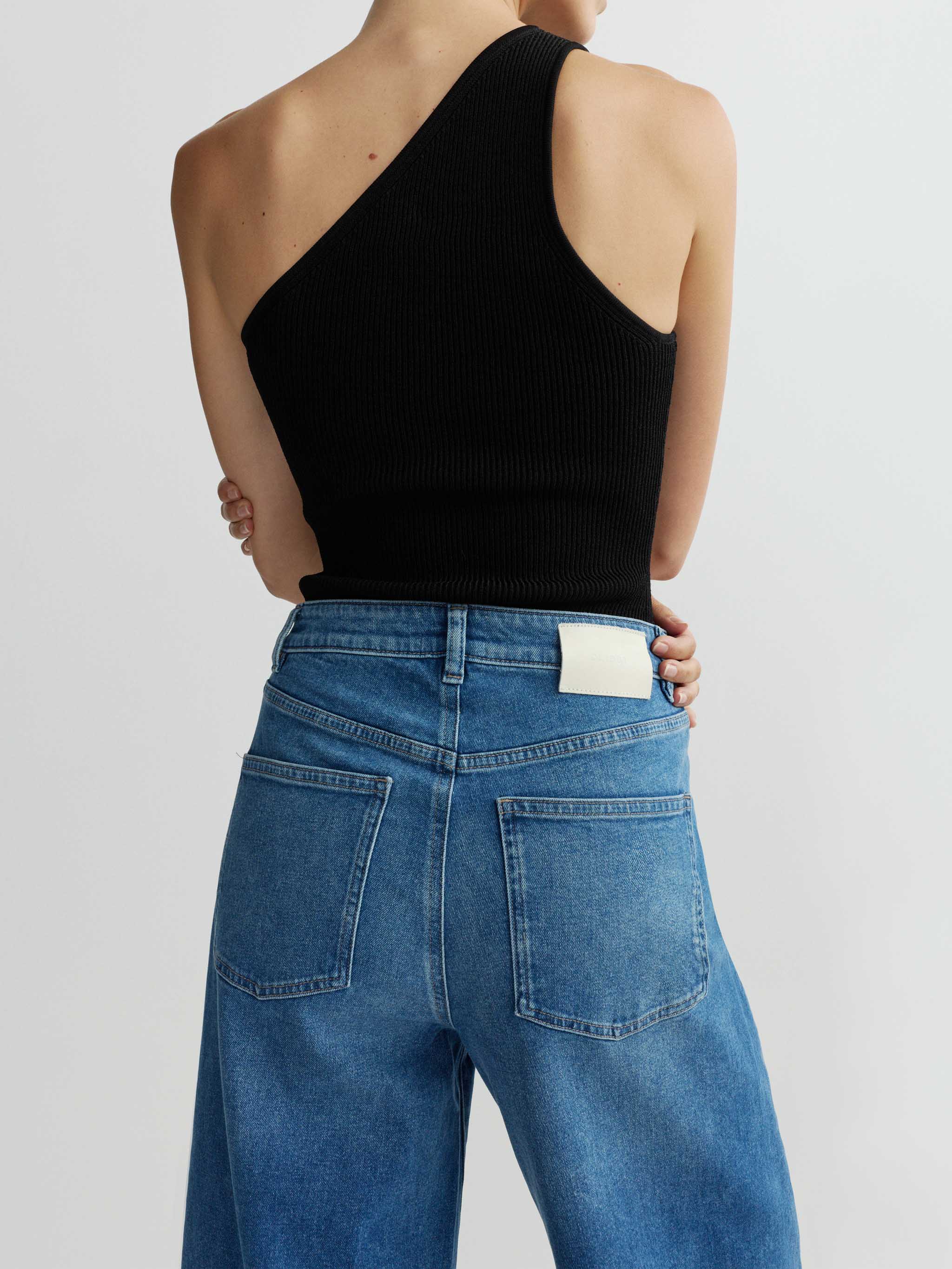 Person wearing a black one-shoulder top and blue jeans on a light gray background