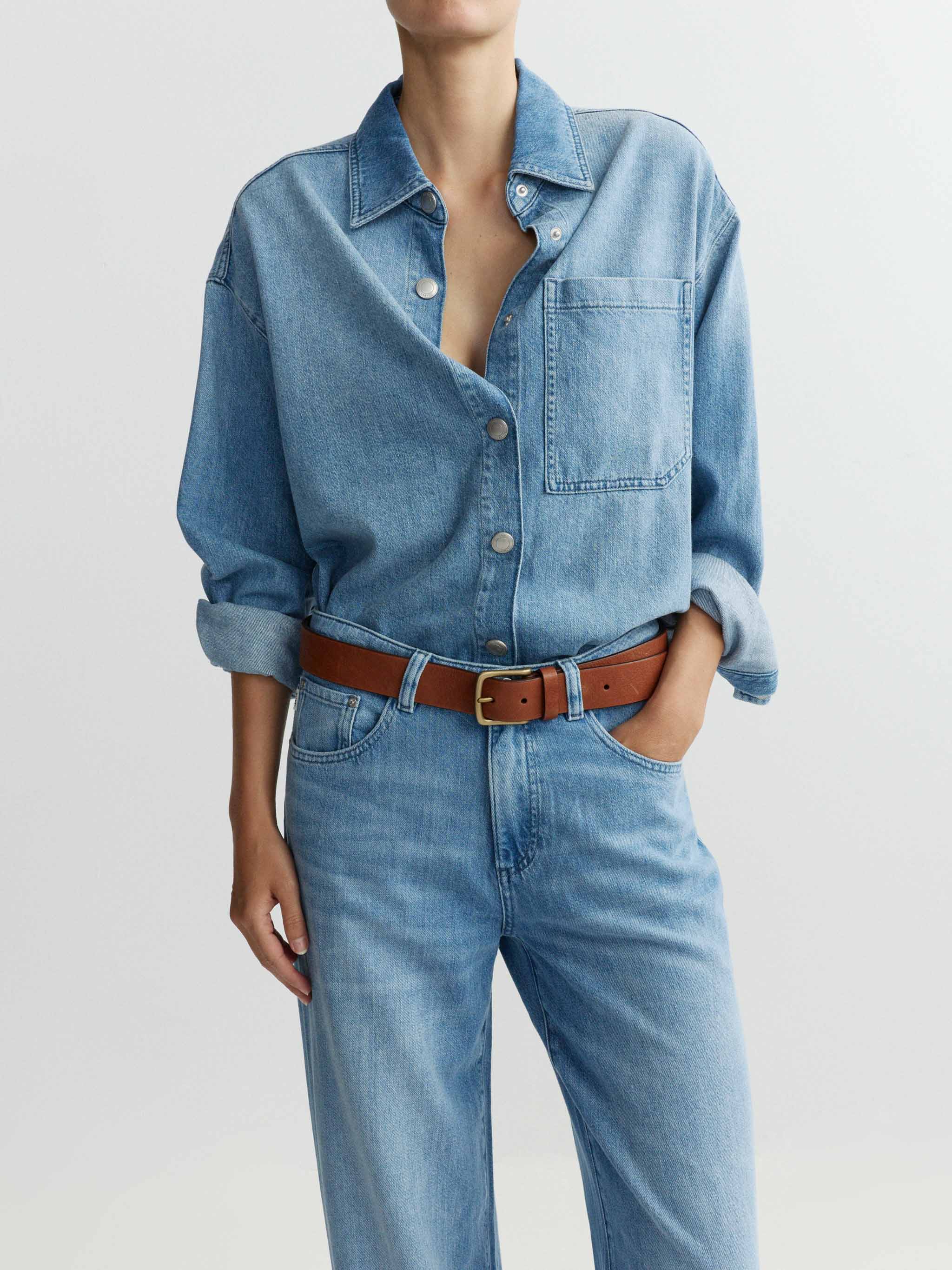 Person wearing a denim outfit with a brown belt on a light gray background