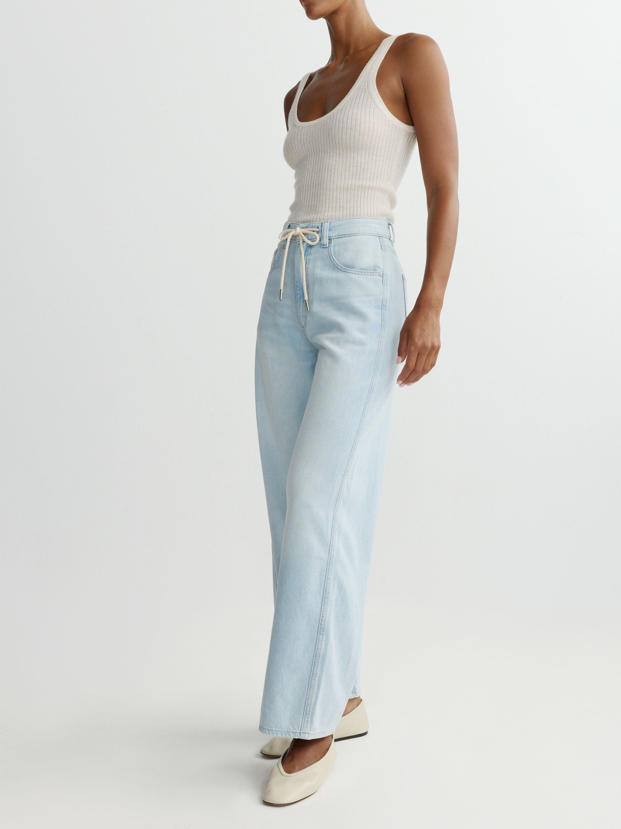 Woman wearing light blue jeans and a white tank top on a plain background