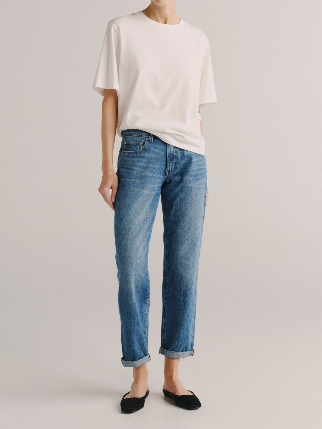 A model wearing a white oversized t-shirt, blue cropped jeans with rolled cuffs, and black flat shoes, standing against a light gray studio background.
