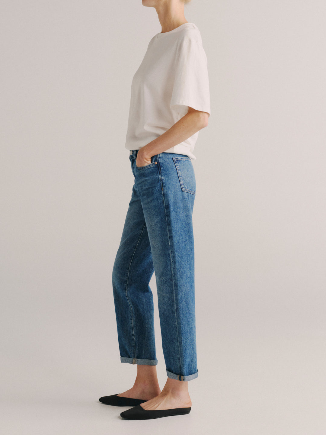 A model shown from the side wearing blue wide-leg cropped jeans with rolled cuffs, a white short-sleeve top, and black flat shoes against a light gray studio background.