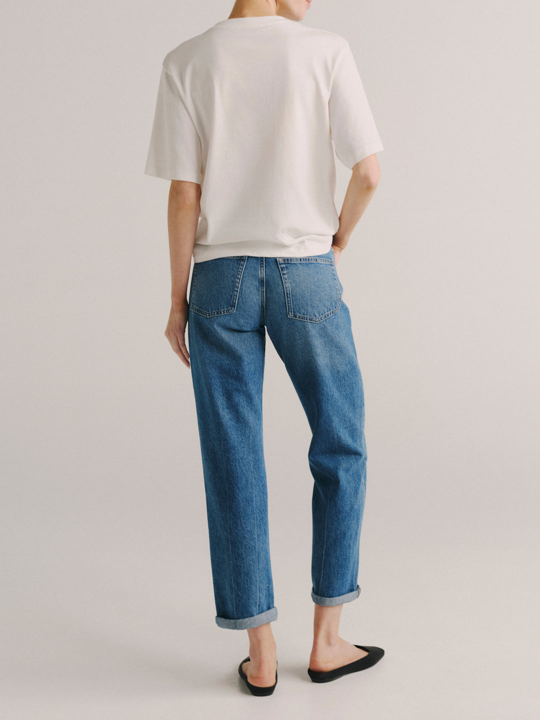A model shown from the back wearing blue straight-leg jeans with rolled cuffs, a white t-shirt, and black flat shoes, set against a plain light gray studio background.
