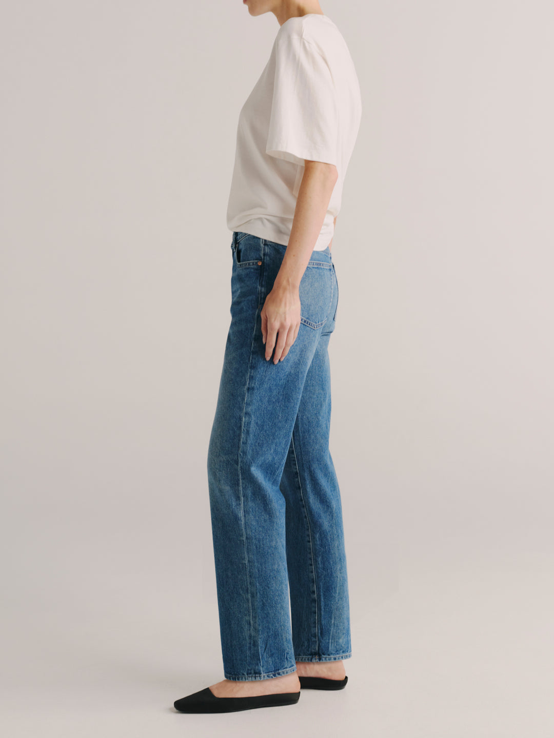A side-profile view of a model wearing blue wide-leg jeans, a white short-sleeve top, and black flats against a light gray studio background.