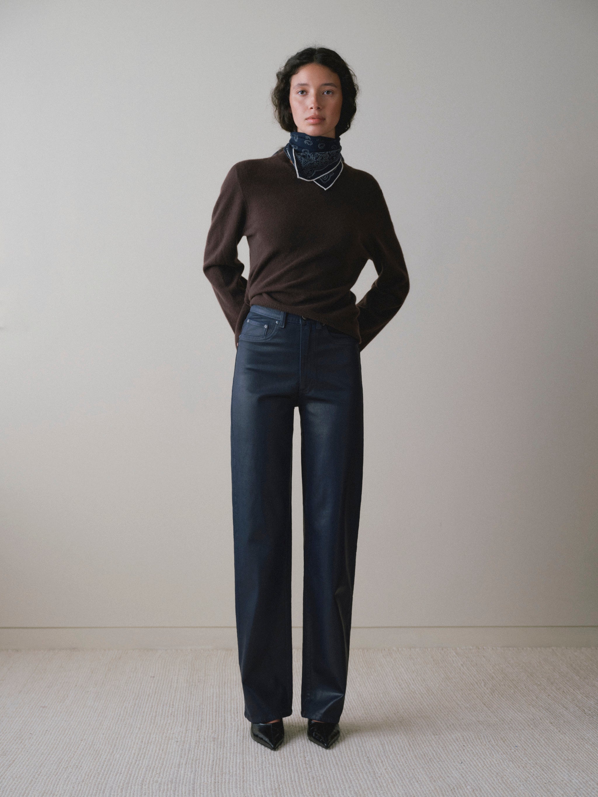 DL1961 women's Karmine Refined Wide Leg High-Rise jeans in coated navy 'Navy Blue Coated', front view with a brown V-neck knit and neck scarf.
