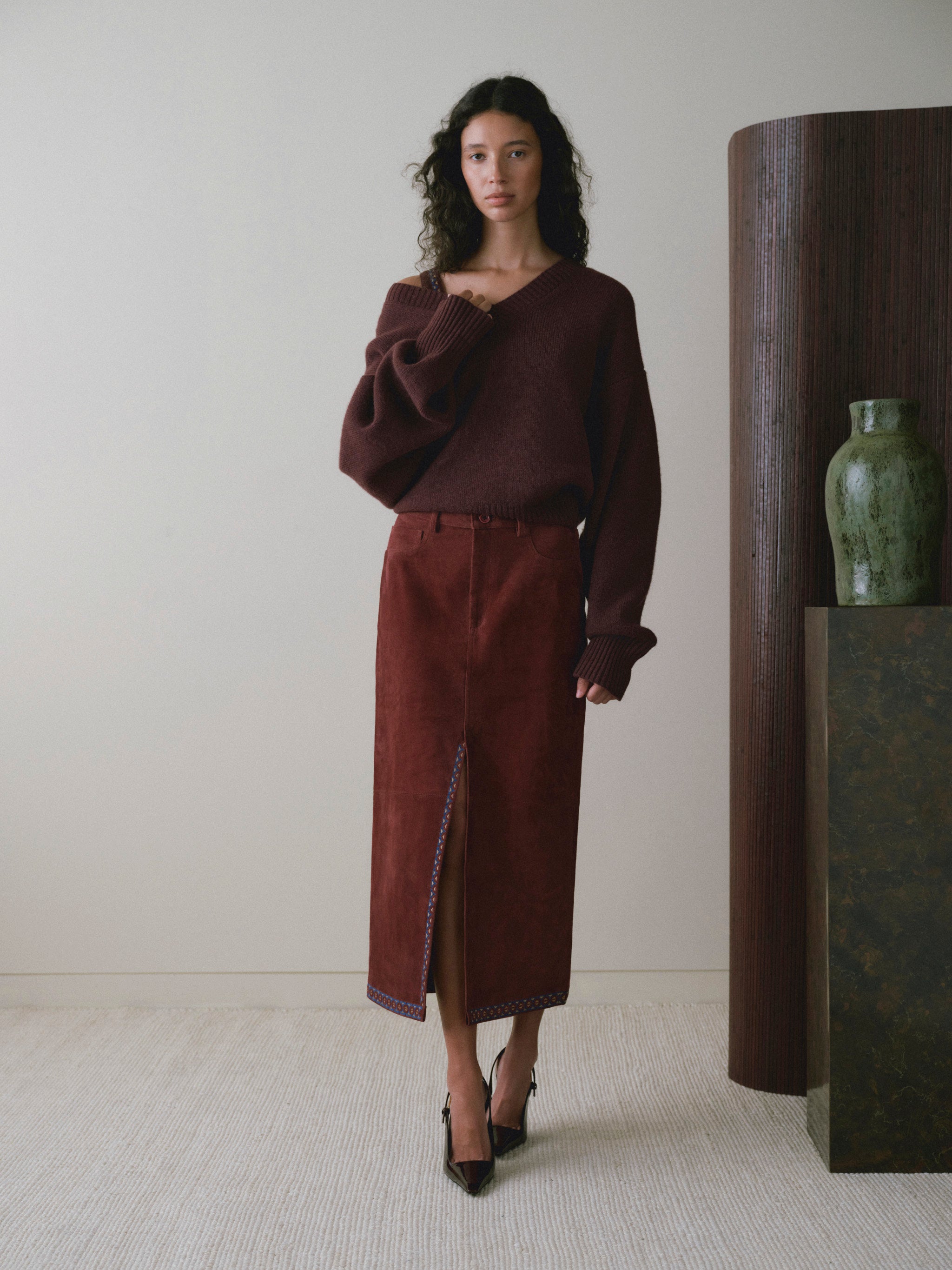 DL1961 women's Robbie oversized V-neck sweater in bordeaux, full-length view worn off the shoulder with a burgundy suede midi skirt.