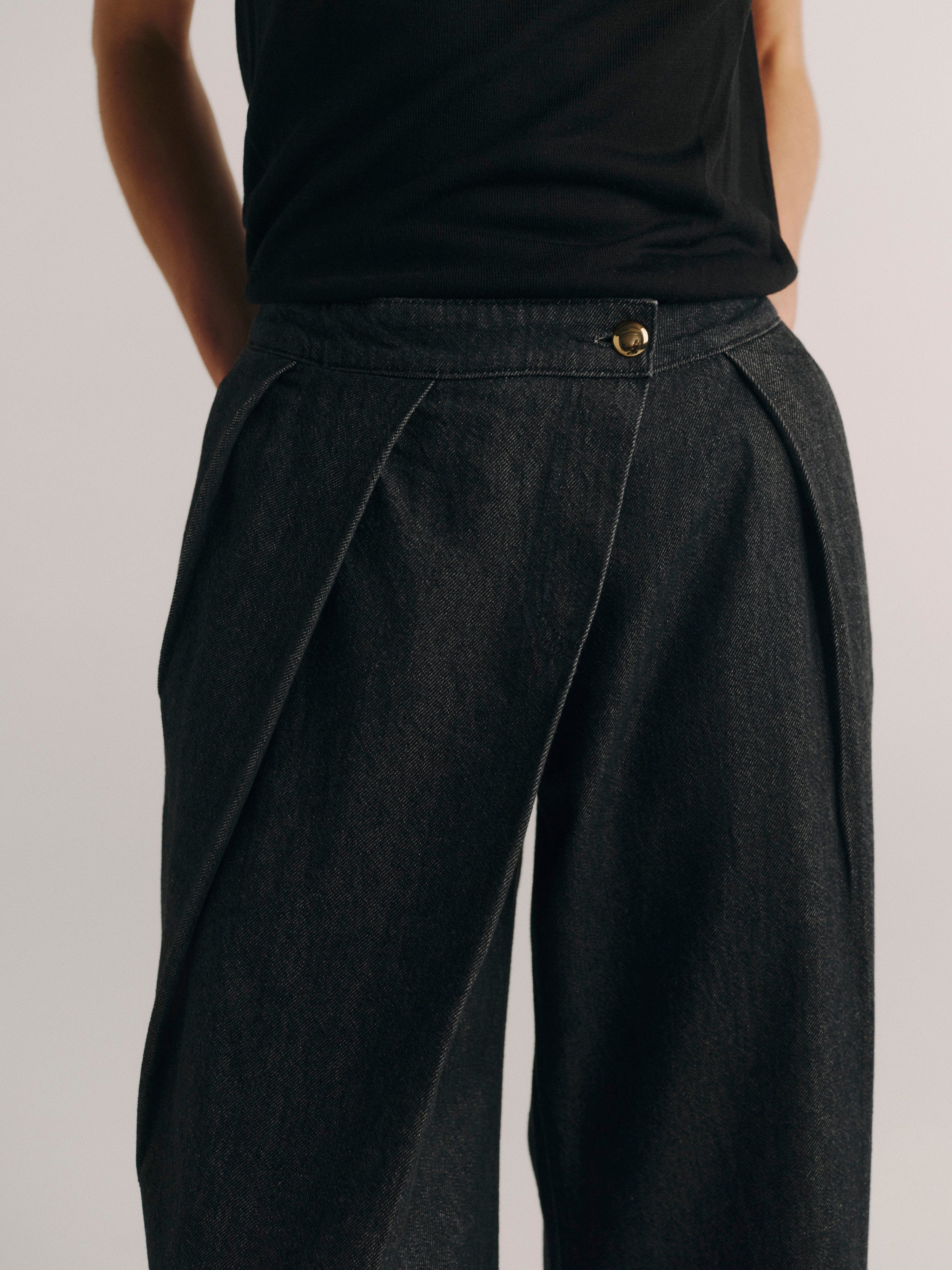 Close-up of a person wearing dark charcoal pleated wide-leg trousers with a black sleeveless top, shown from the back against a light gray studio background.