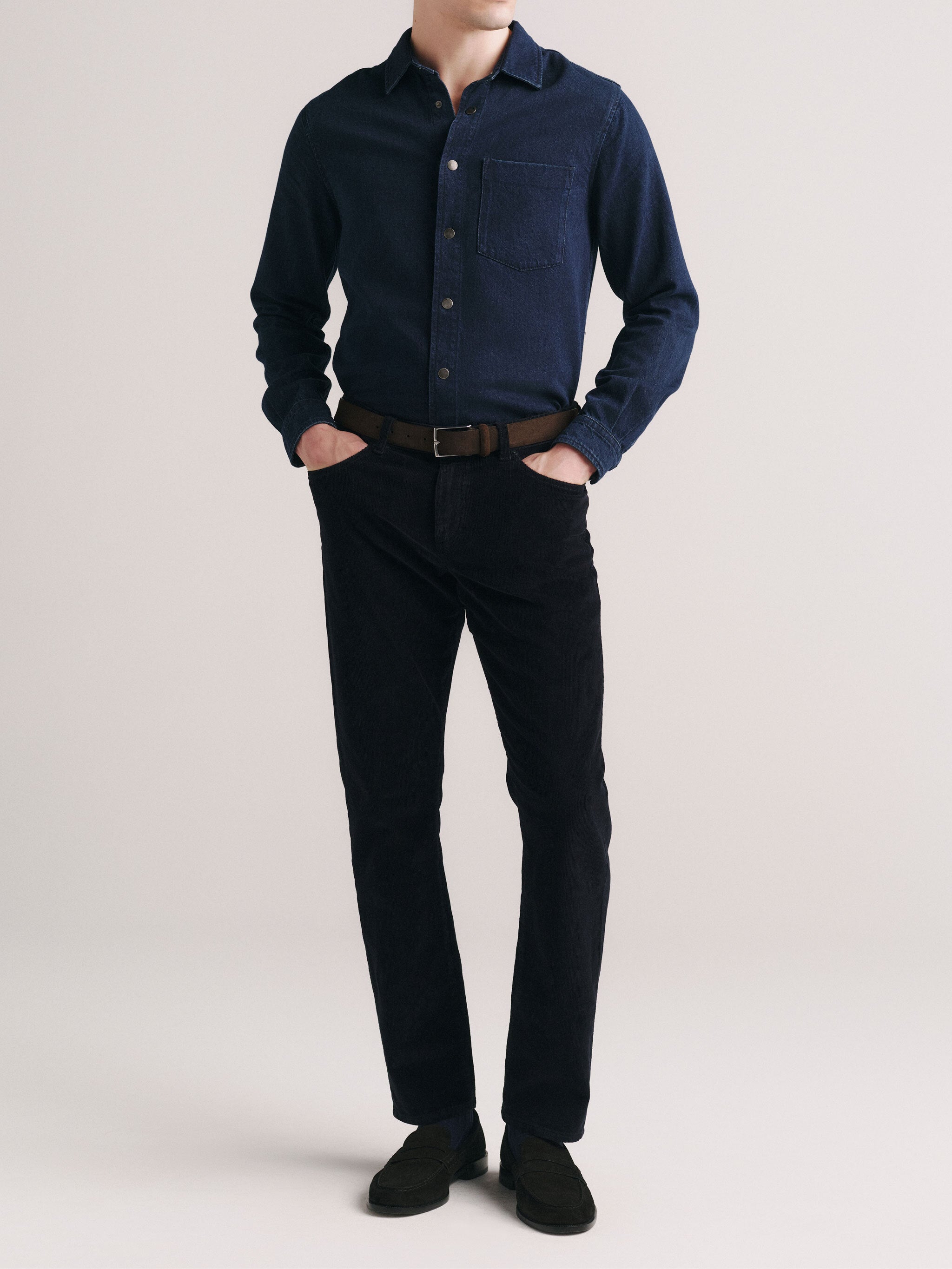 A man wearing a dark navy blue long-sleeve button-up shirt, dark charcoal pants, and black shoes, standing against a light gray studio background.