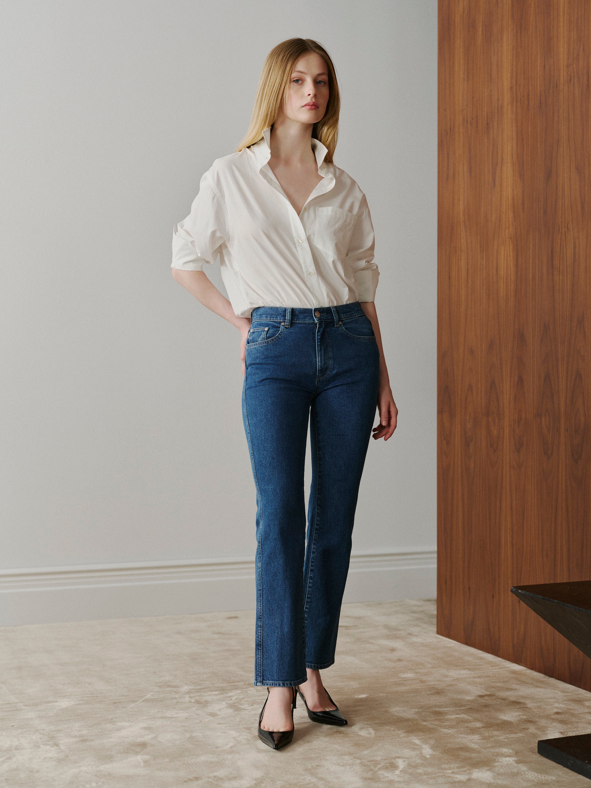 Woman wearing a white shirt and blue jeans standing in a room with wooden paneling.