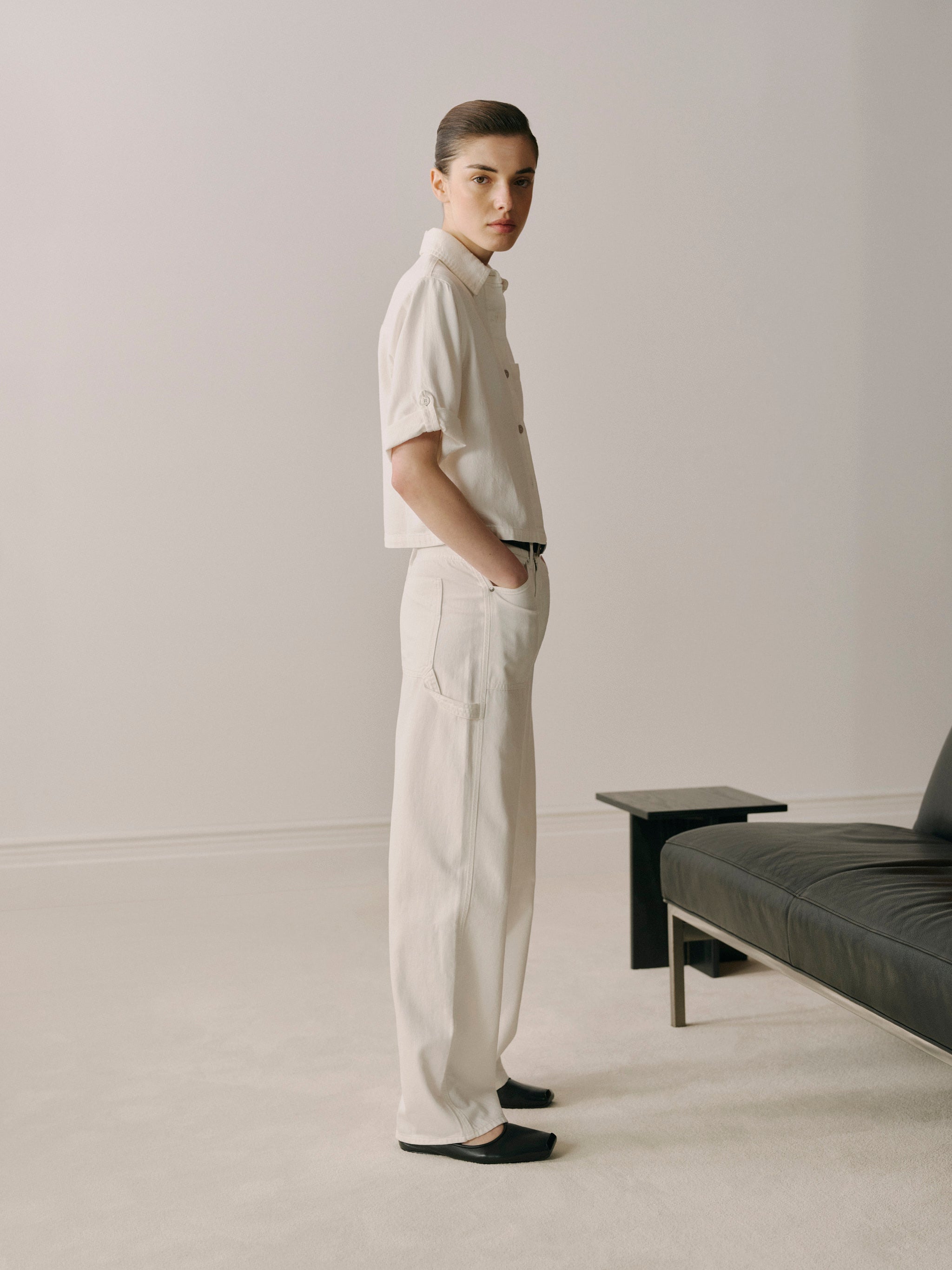 Person wearing a light-colored shirt and pants standing in a minimalistic room with a gray sofa and black table.