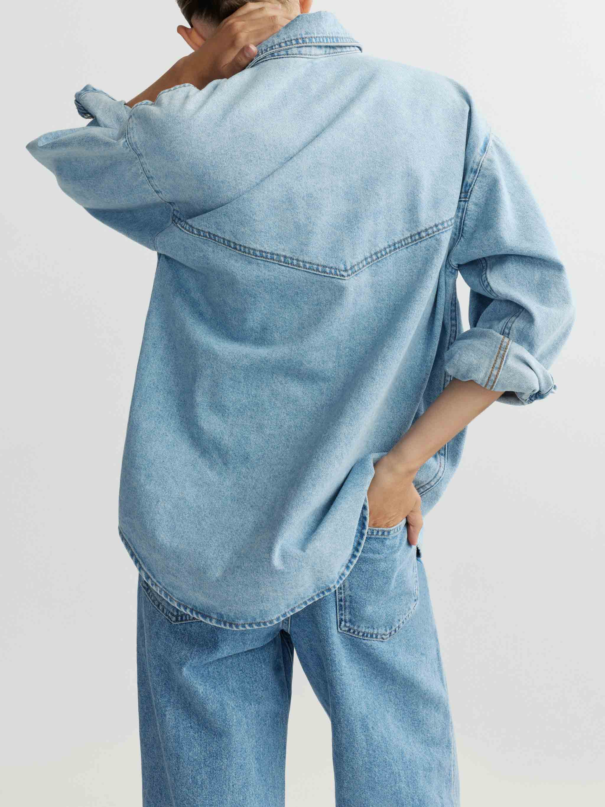 Person wearing an oversized light blue denim jacket and jeans on a plain background