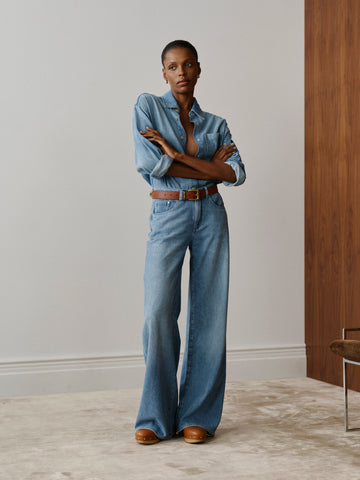 Person wearing a blue denim outfit with a belt, standing in a room with wooden flooring and a neutral wall.