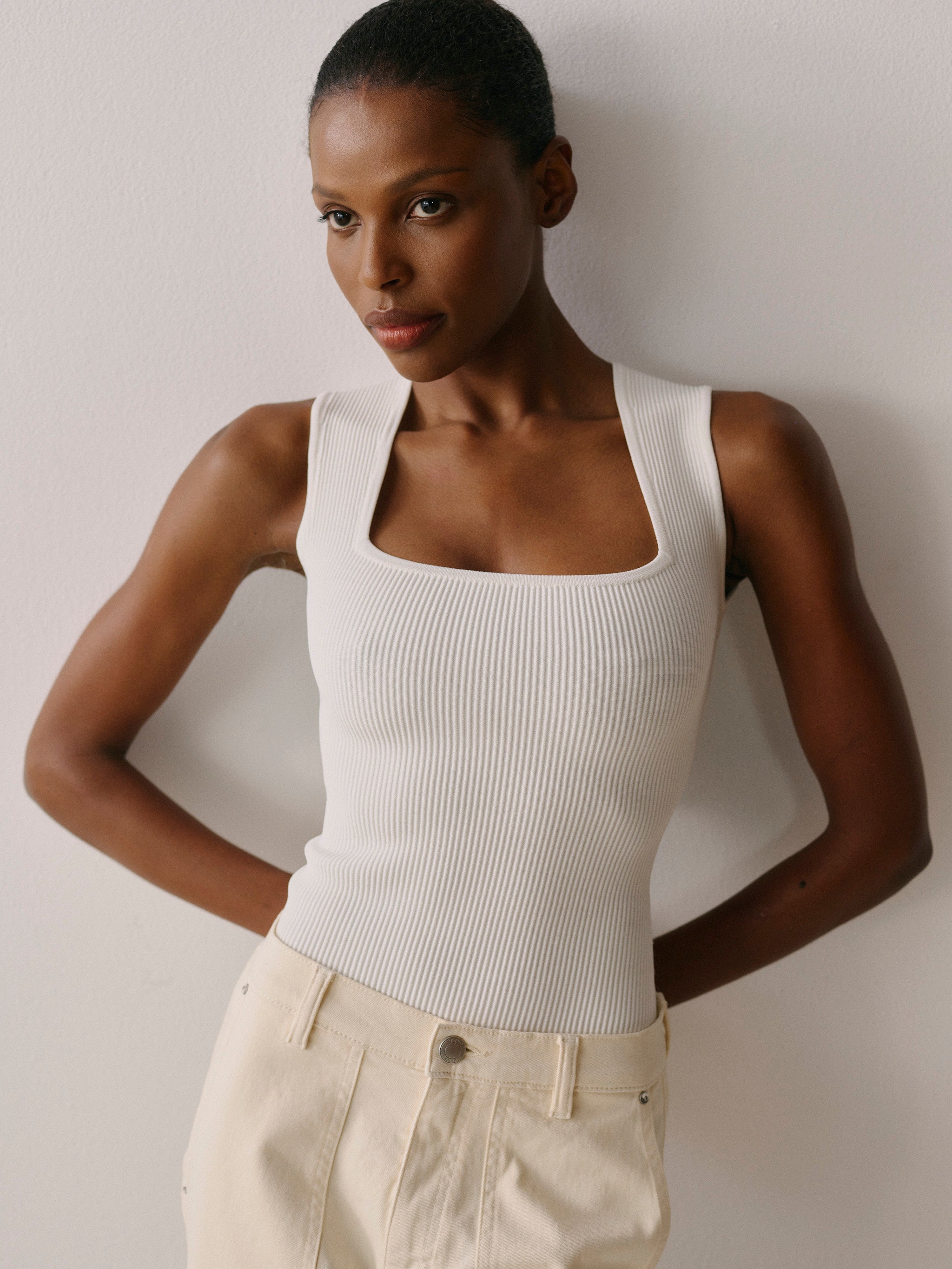 Woman wearing a white tank top and beige pants against a plain background