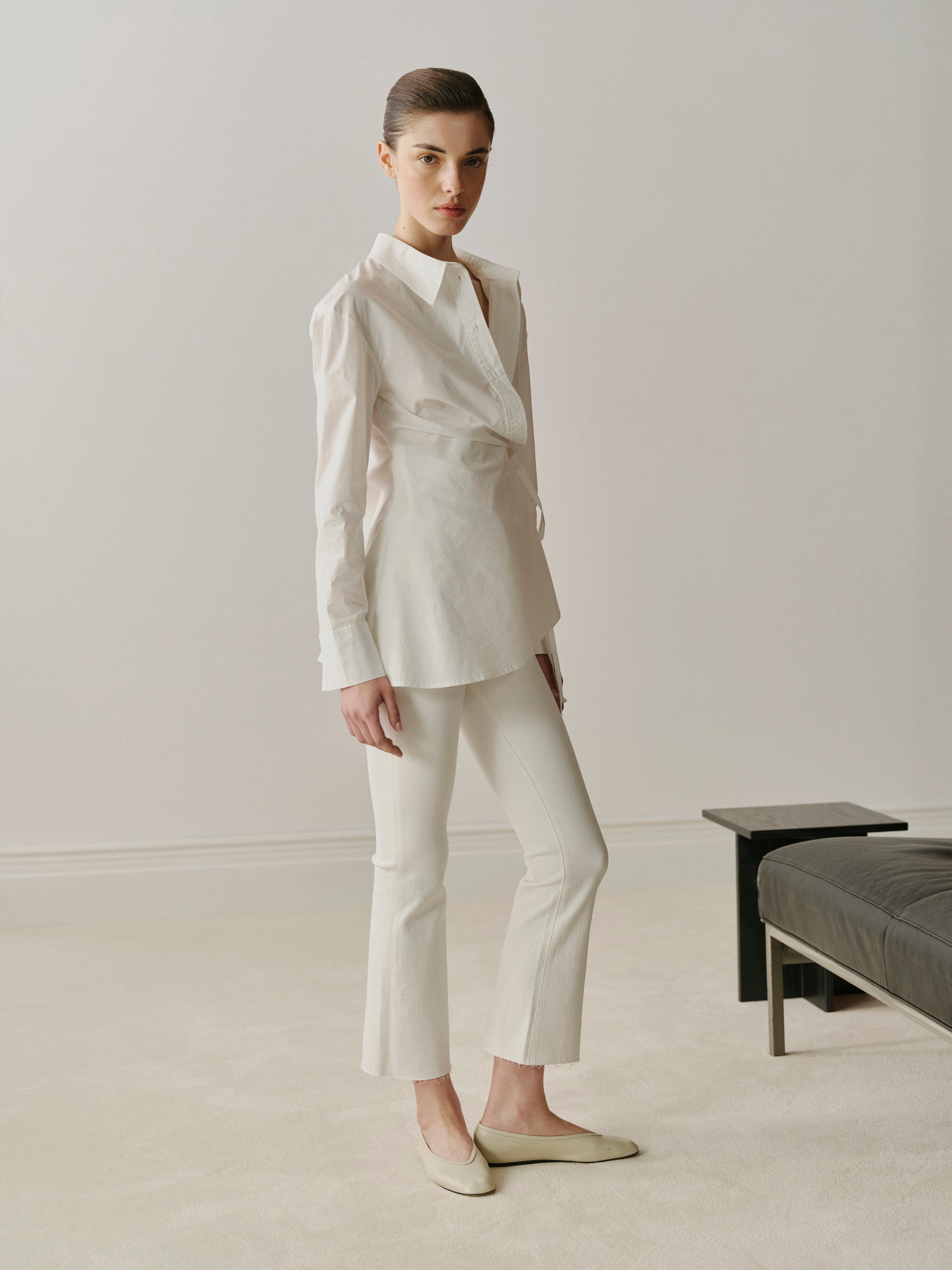 Woman wearing a white suit standing in a minimalistic room with a bench.