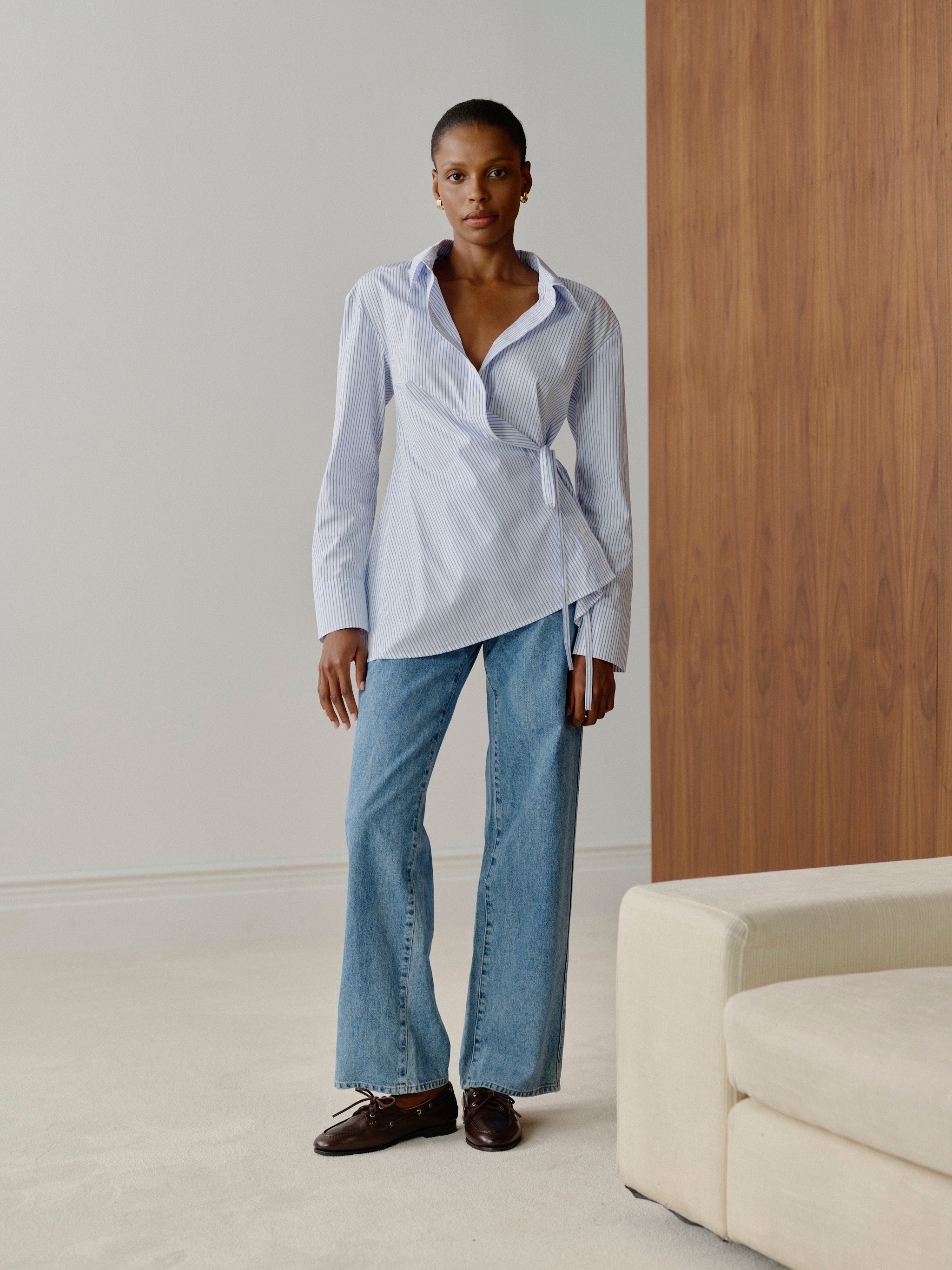 Woman wearing a light blue shirt and jeans standing in a minimalistic room with a wooden wall and white floor.